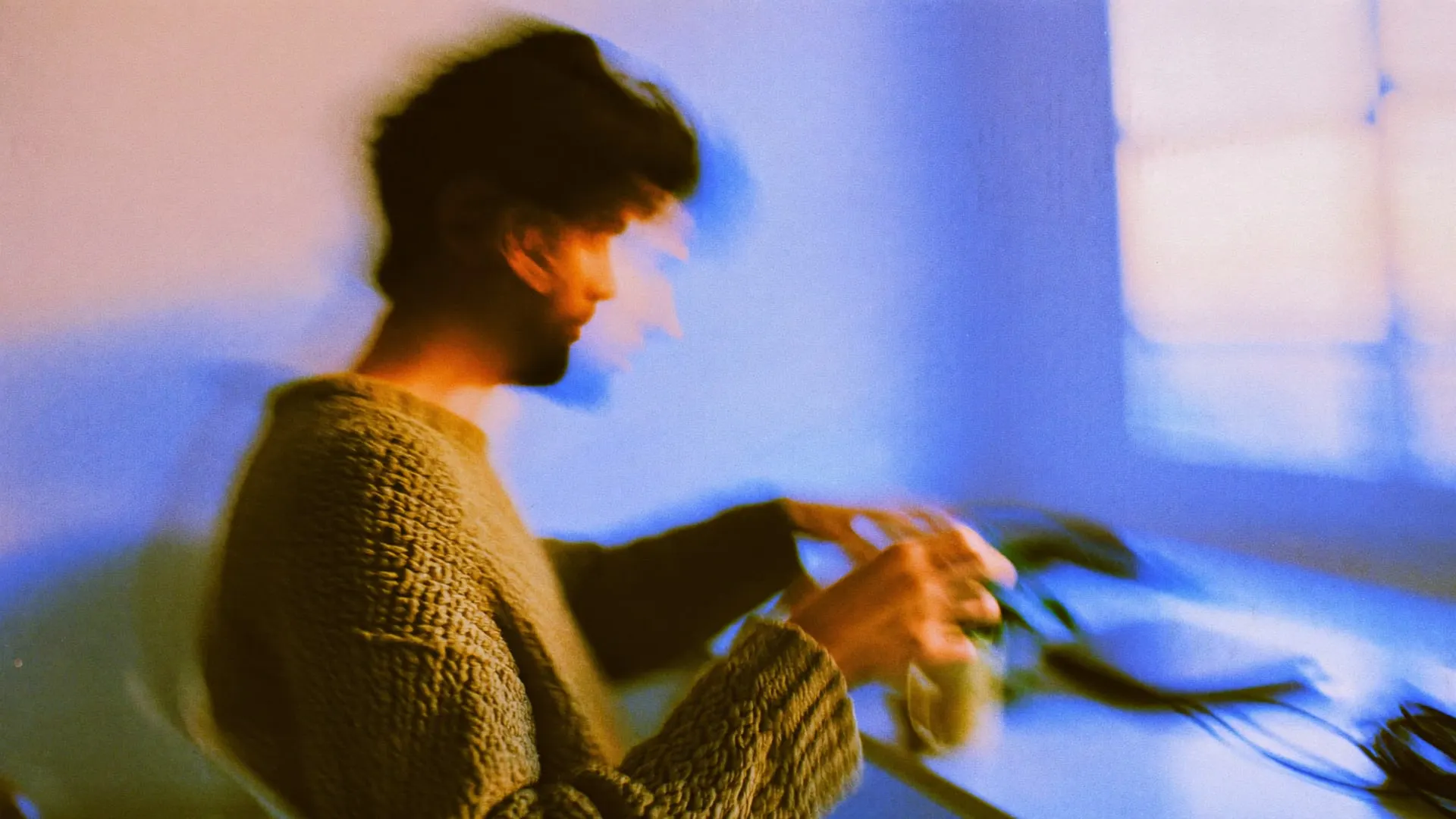 A man at a desk in early morning light adjusting a small plant beside a closed laptop