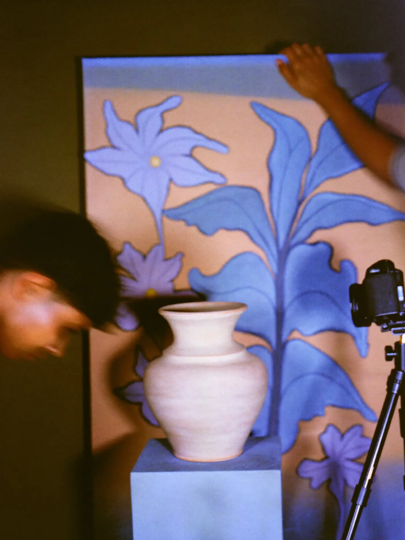 A ceramic vase on a plinth before a painted backdrop, a camera on a tripod beside it