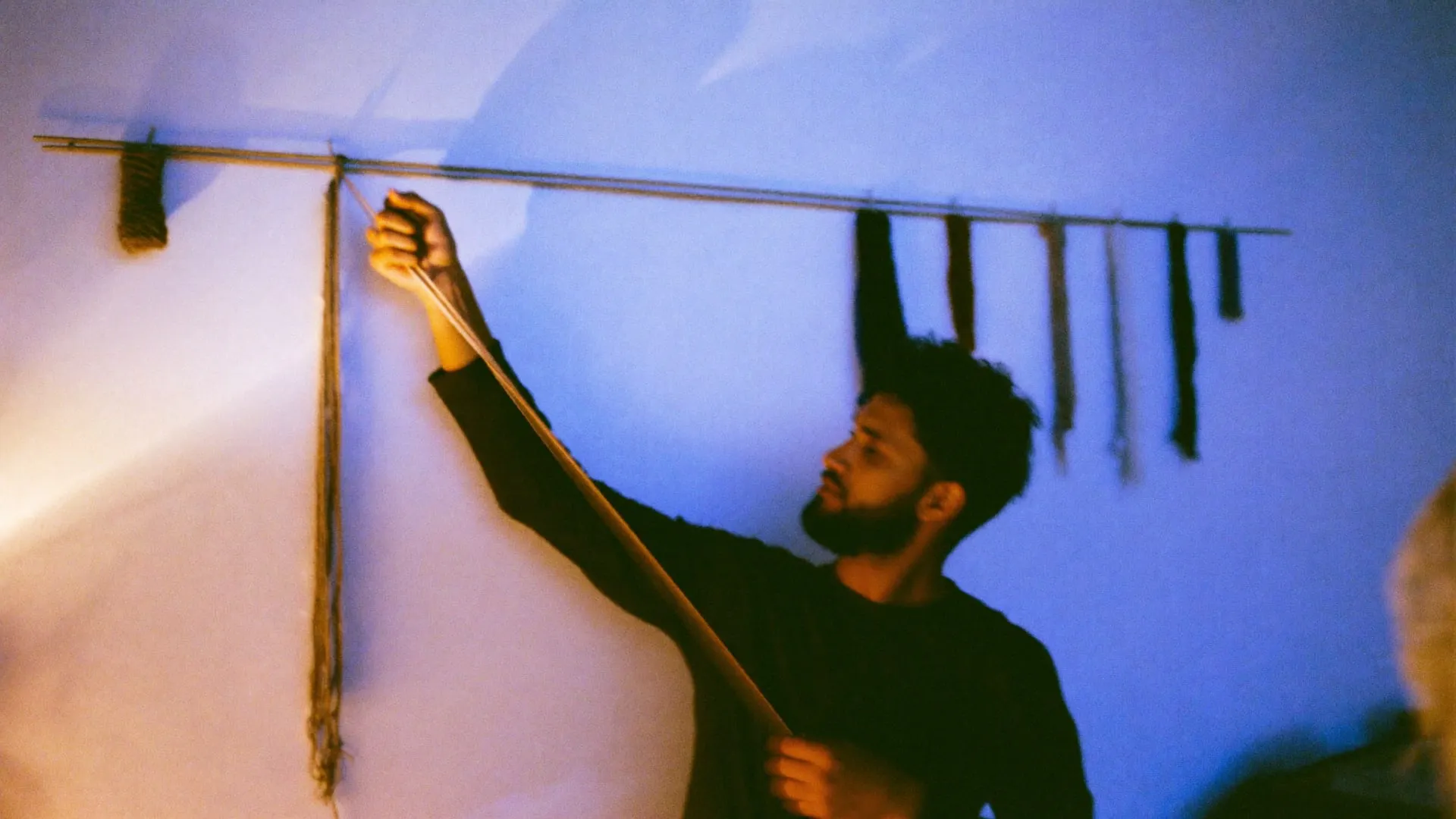 A man stretching coloured yarn between pins along a studio wall at night