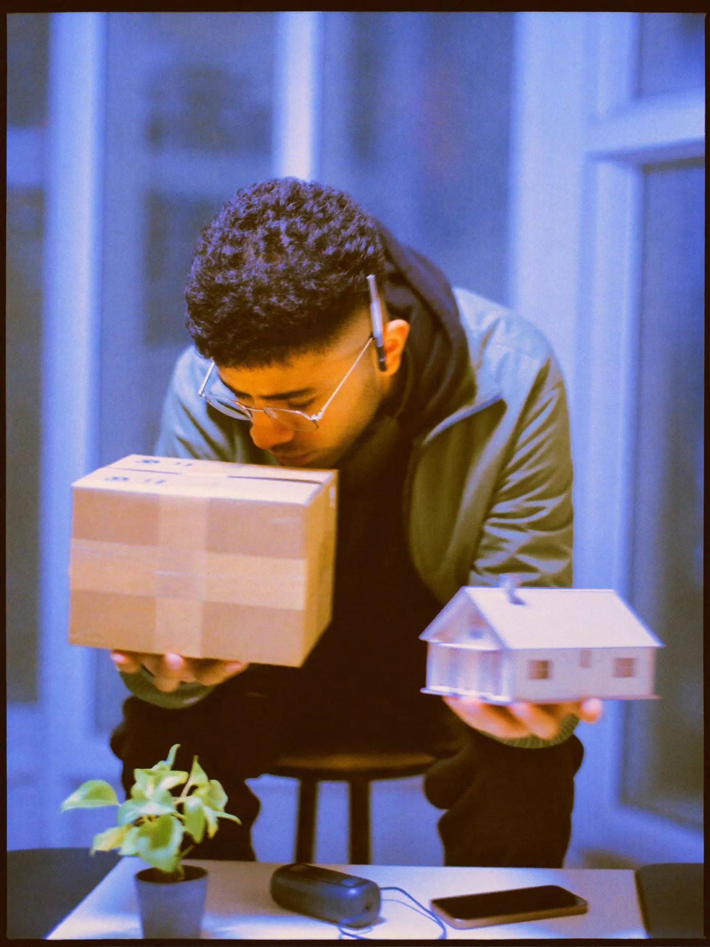 A young man in glasses weighs a delivery box in one hand and a model house in the other