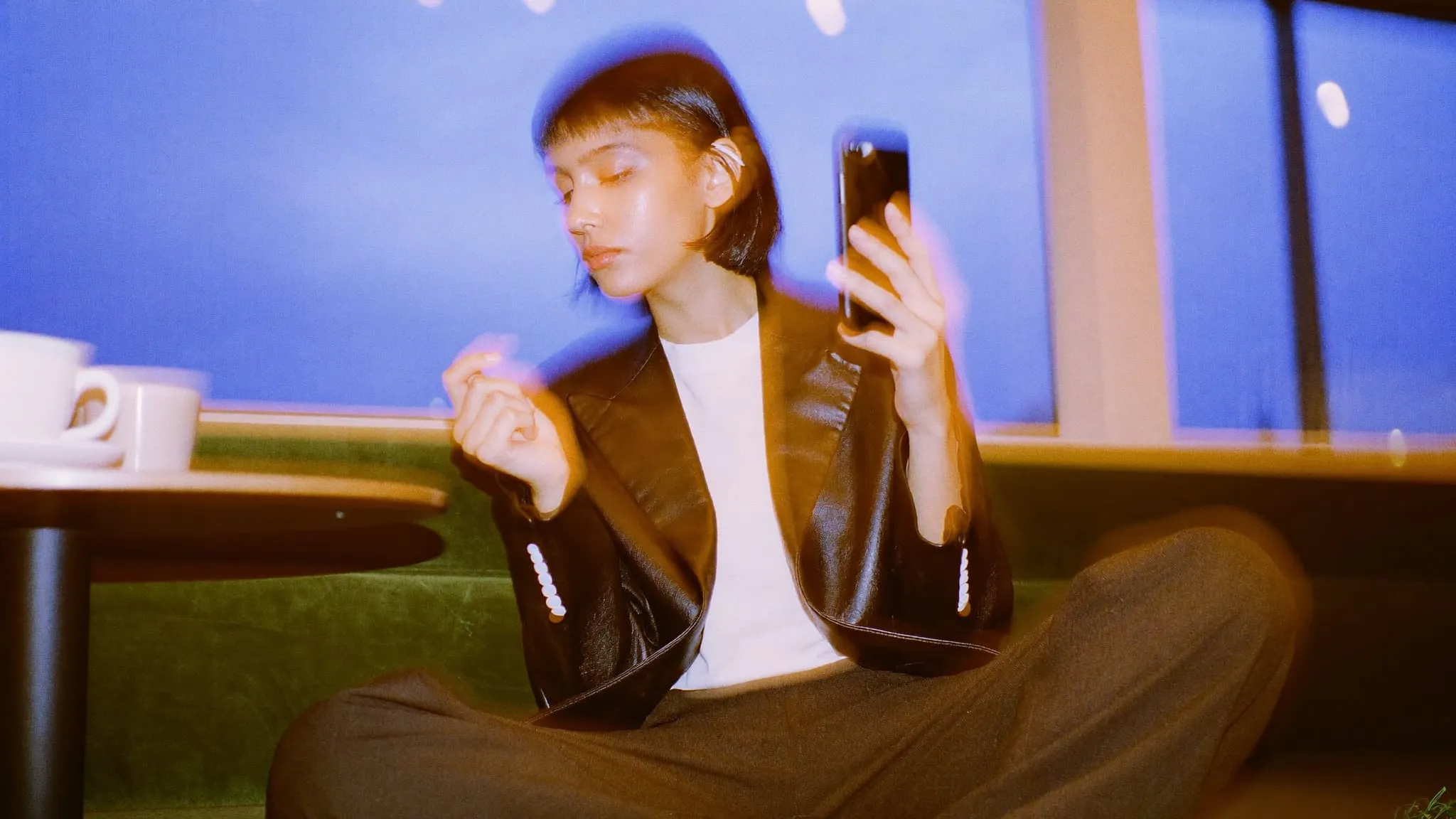 A young woman with a black bob sits cross legged on a green velvet cafe banquette, looking at her phone