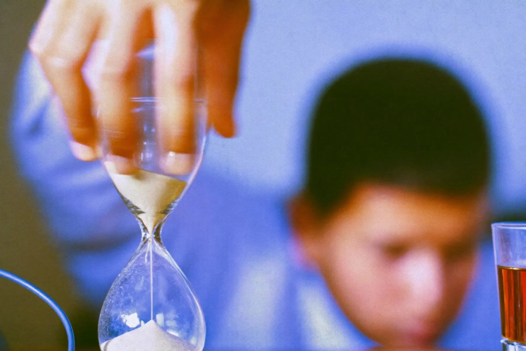 A hand turning over an hourglass on a wooden desk beside a glass of tea