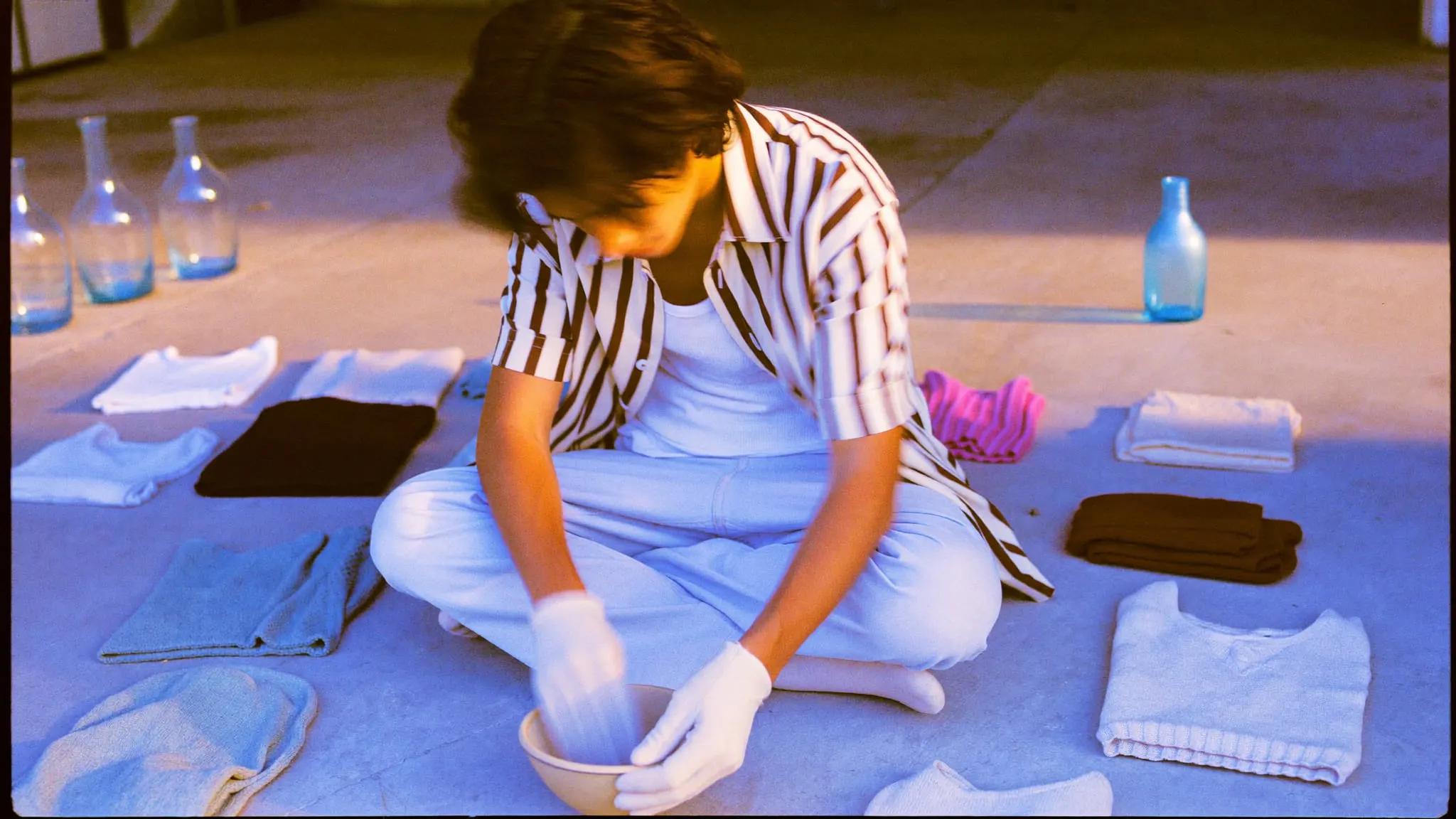 A young man in white gloves holding a ceramic bowl beside rows of folded clothes and ceramics