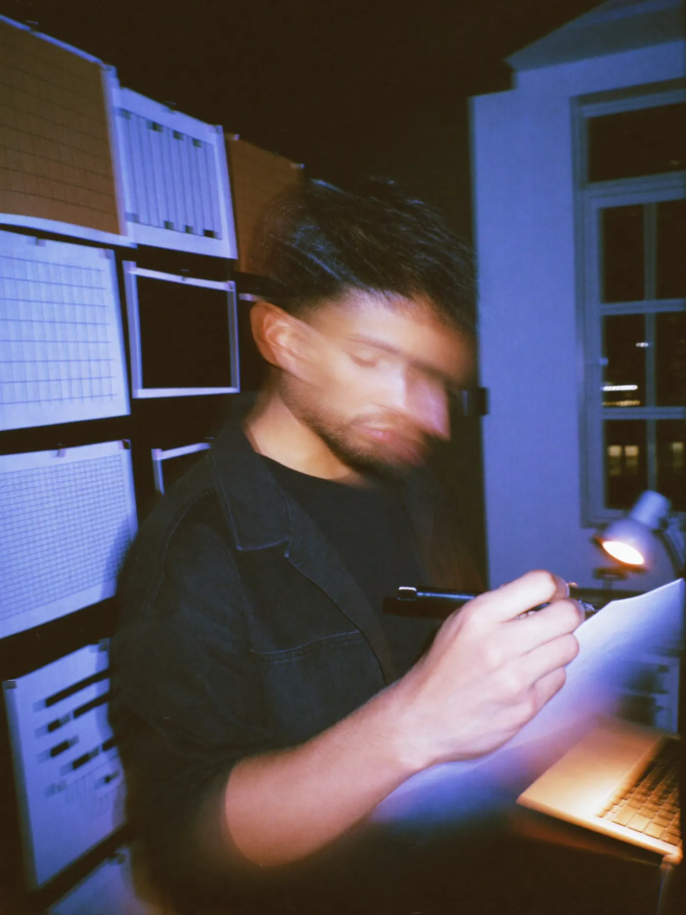 A bearded man in a charcoal jacket marks printed page layouts pinned to a studio wall at night