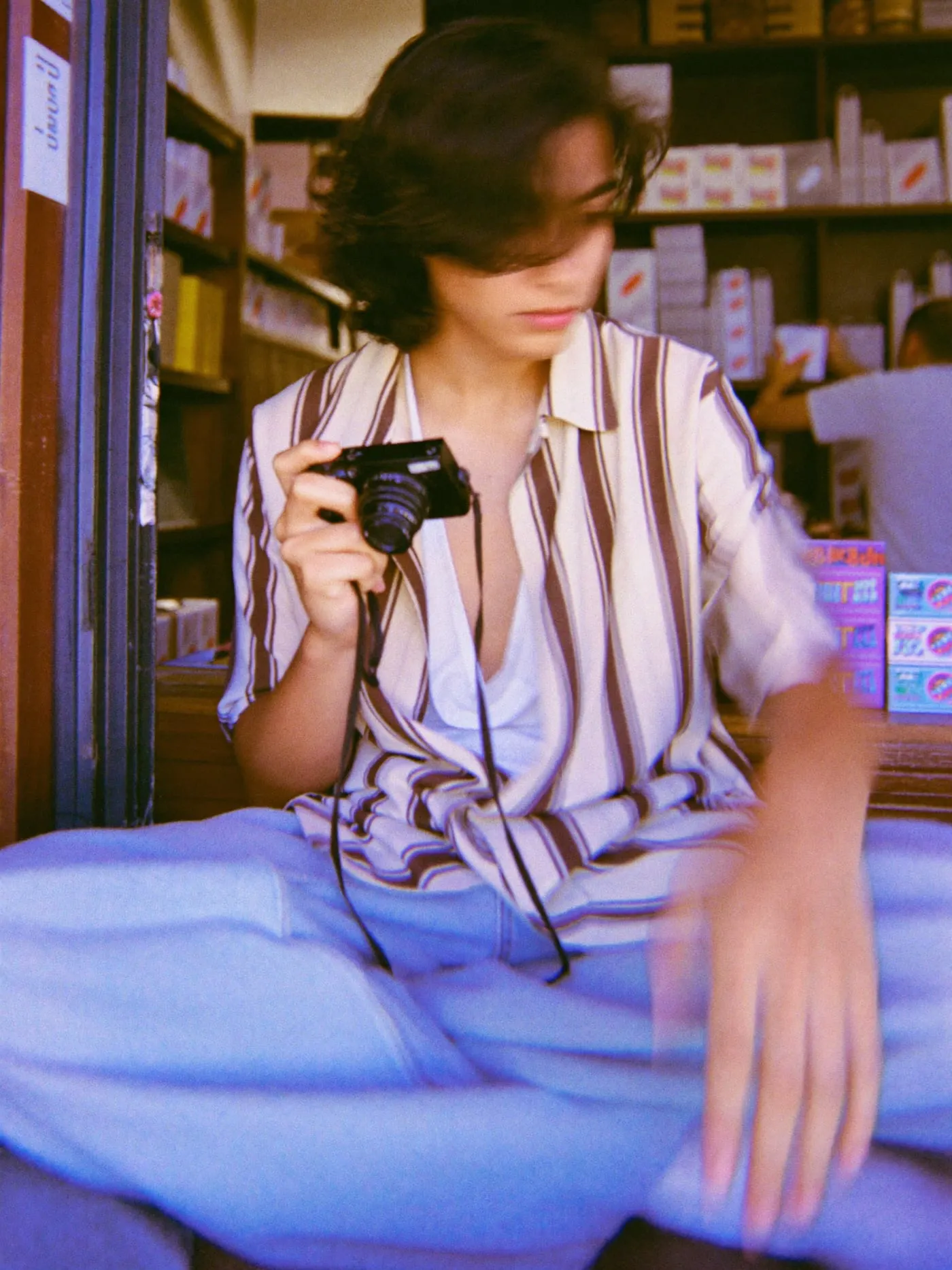 A young man sitting on a shop counter, photographing shelves of boxed goods with a film camera
