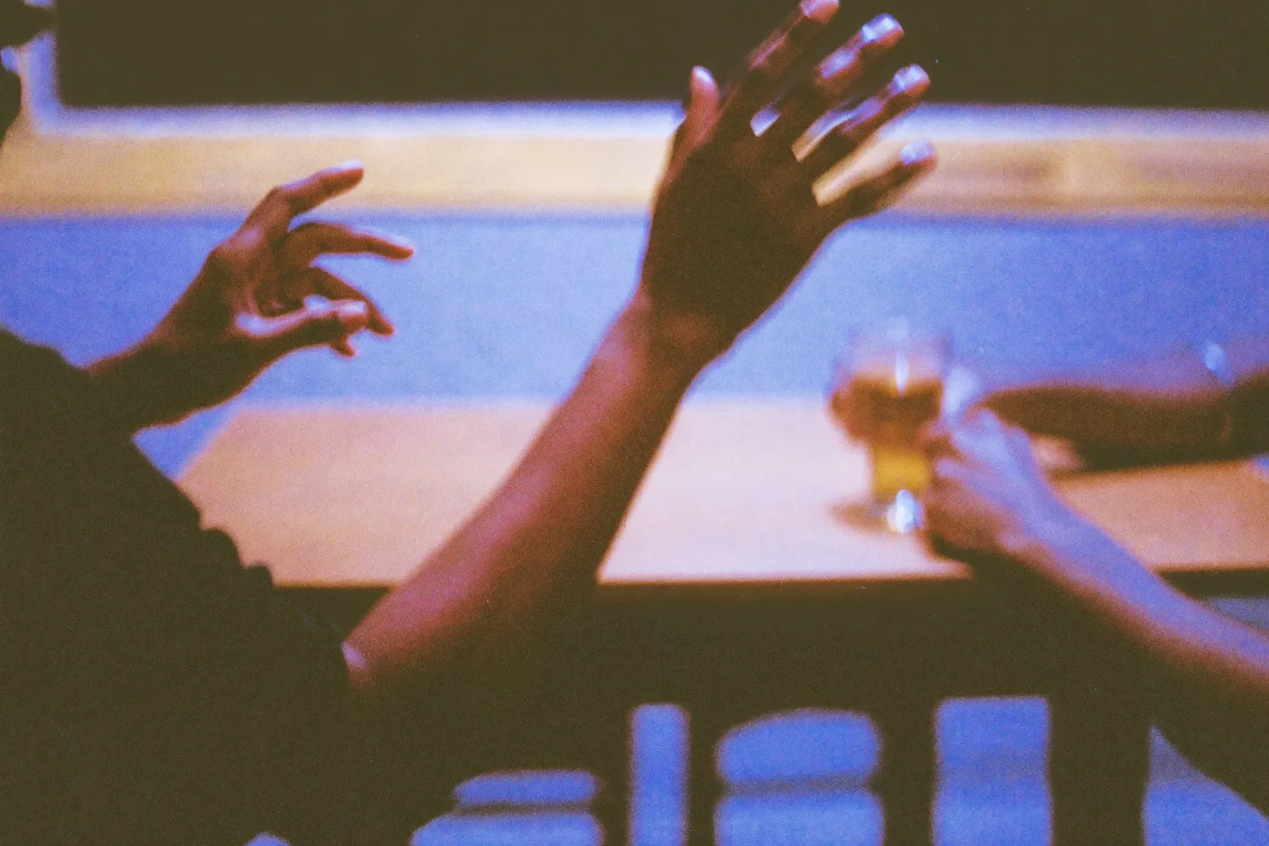 Two colleagues mid-conversation across a table, one pair of hands gesturing, tea and paper between them