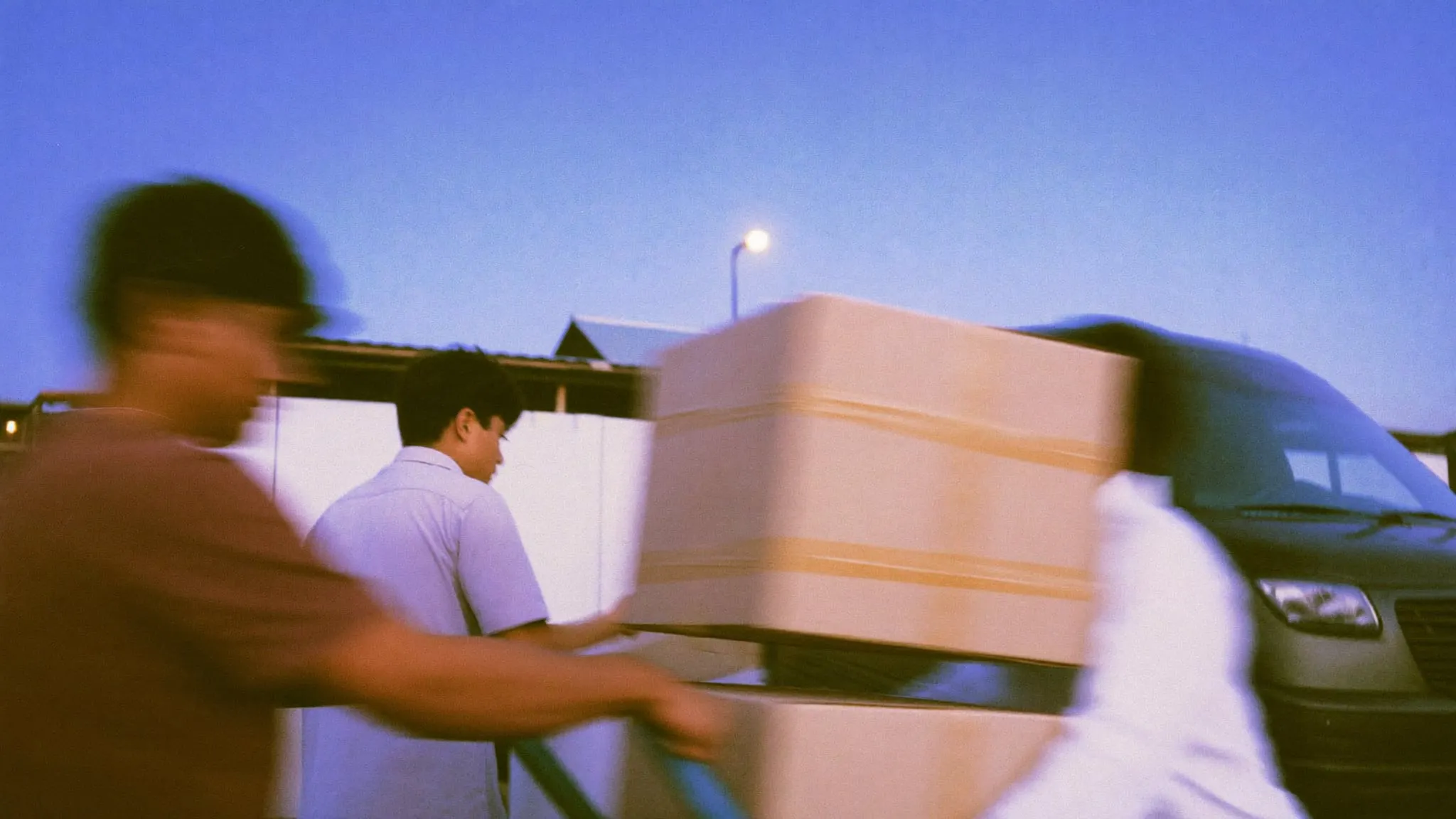 Two workers wheeling a hand truck of cartons toward a van at a loading bay at dawn