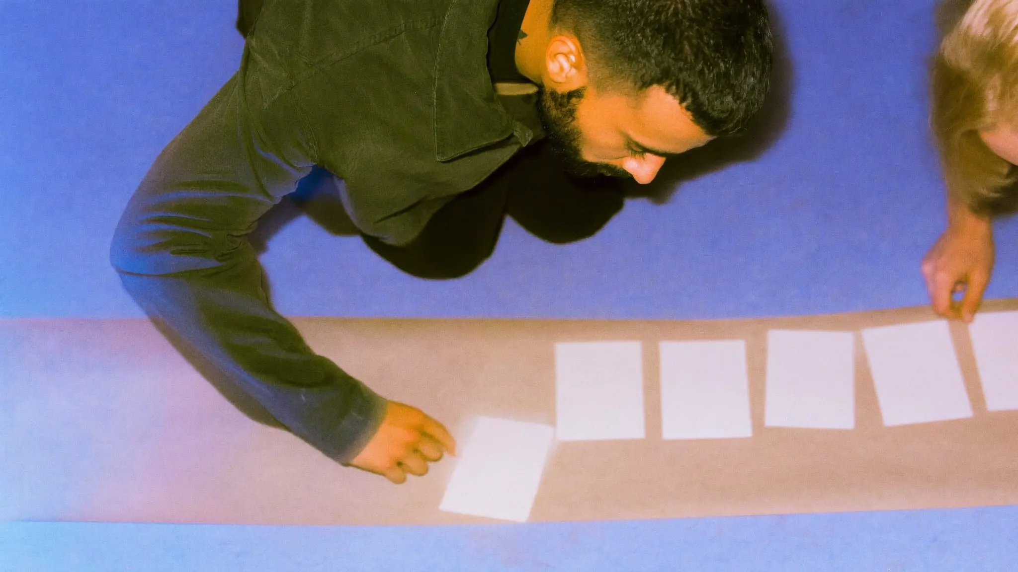 A bearded man crouching on a studio floor laying blank cards in a row along kraft paper with a colleague