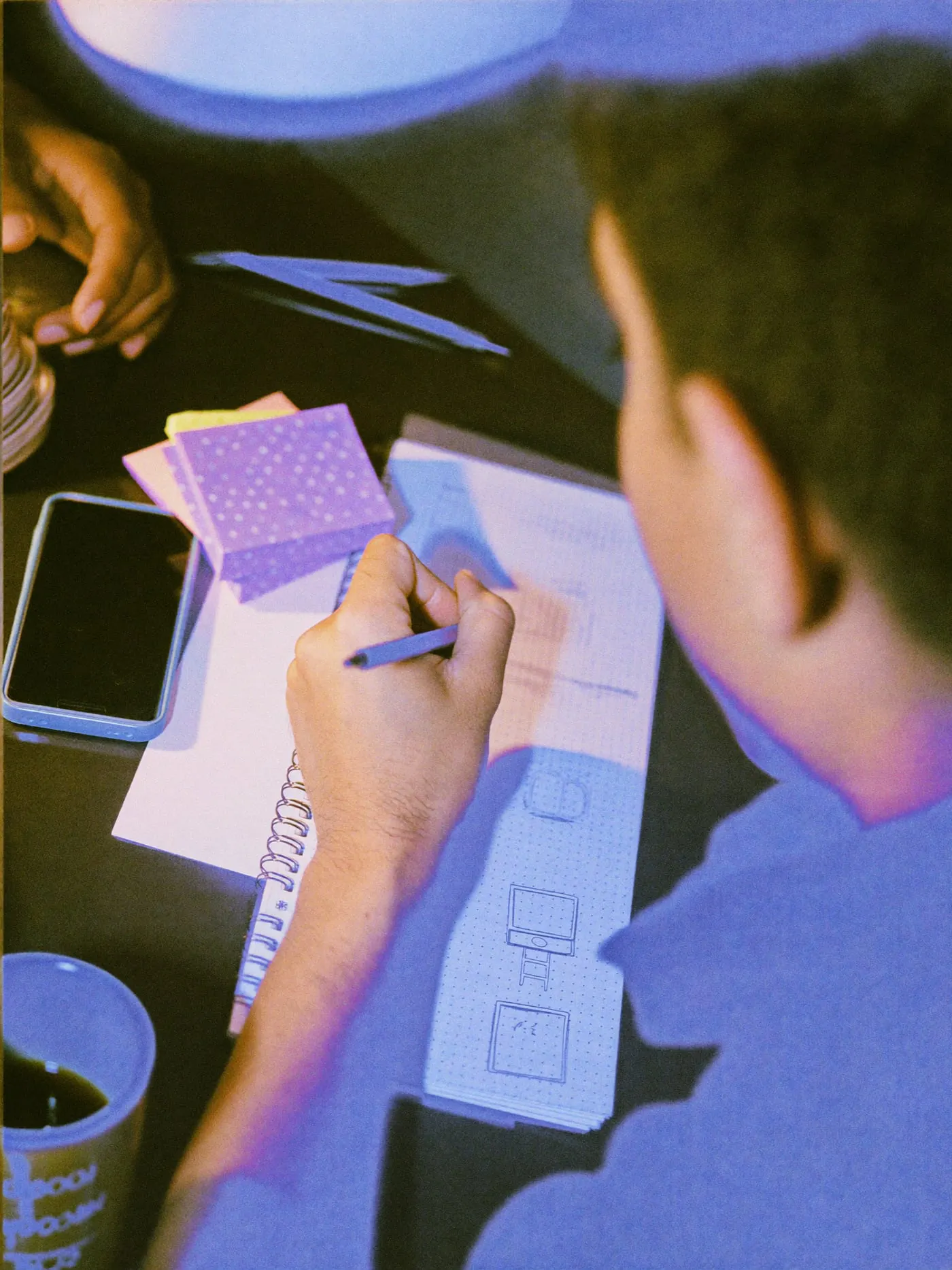 Hands sketching small screen outlines in a dotted notebook beside a face down phone
