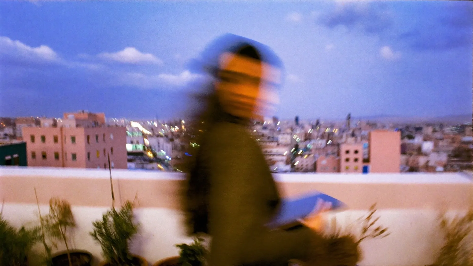 A young woman seen from behind walking across a Cairo rooftop terrace at blue hour with a closed laptop
