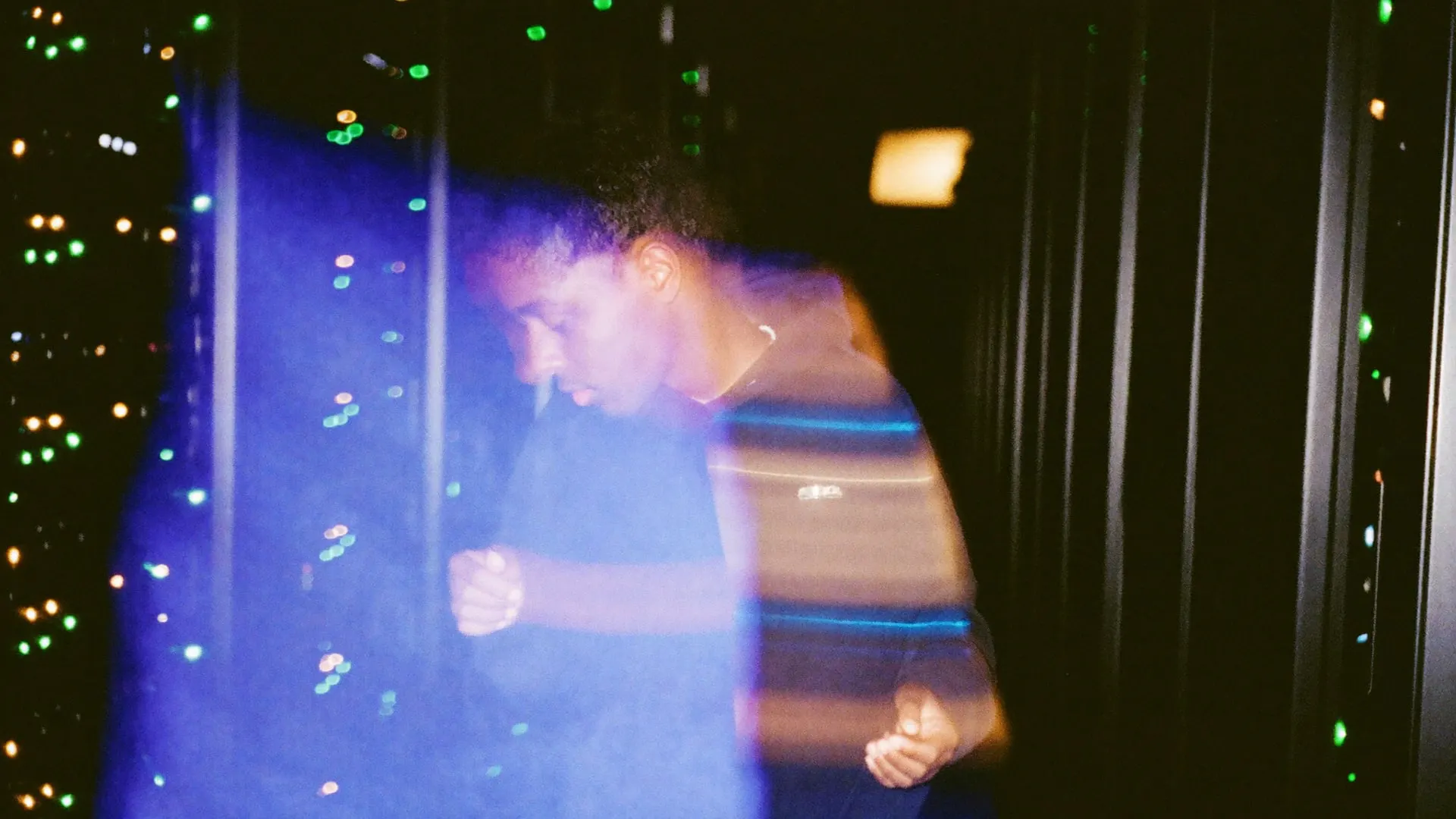 A young engineer walking fast down an aisle of server racks, status lights stretched into long lines.