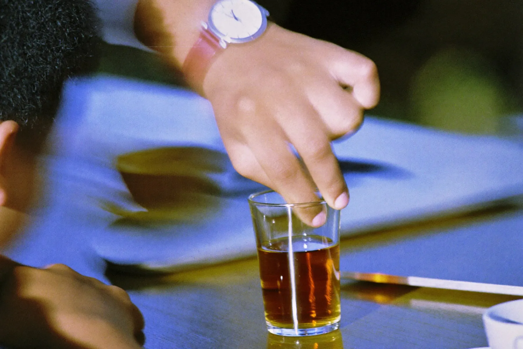 A hand adjusting an analogue wristwatch on a desk beside a closed laptop and a glass of tea