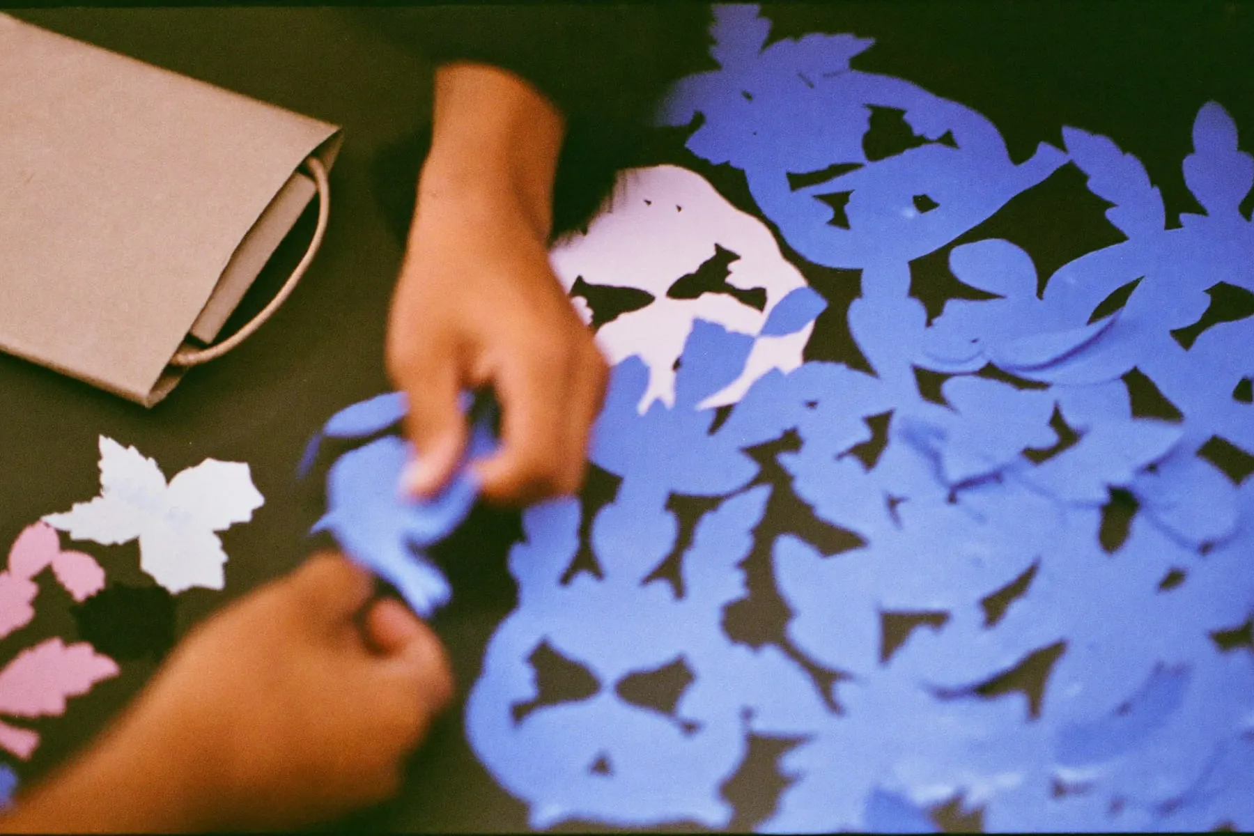 Hands arranging cut paper shapes, fabric swatches and a plain kraft bag on a dark table