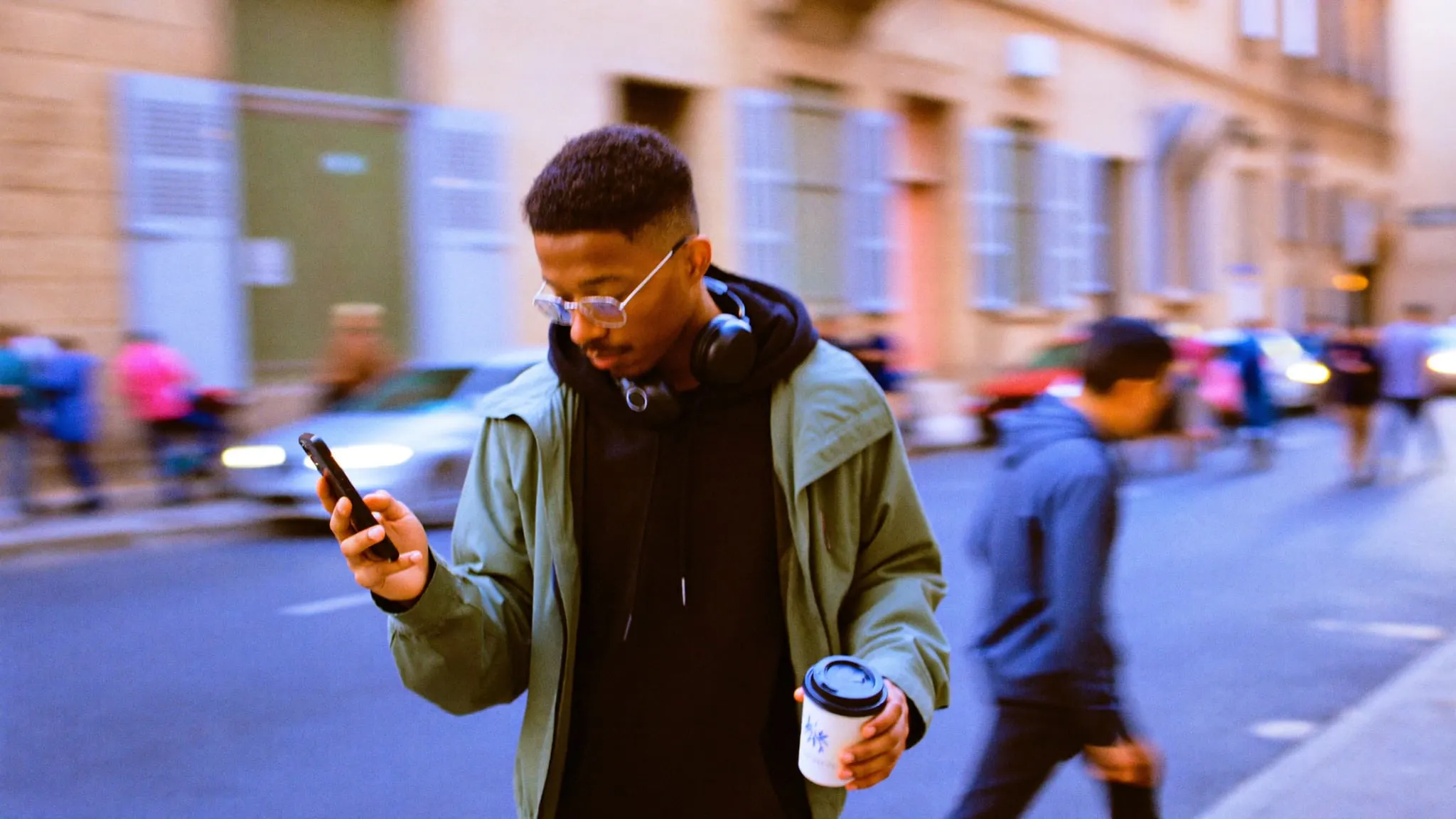 A young man in glasses crossing a Downtown Cairo street with a coffee, using his phone one handed