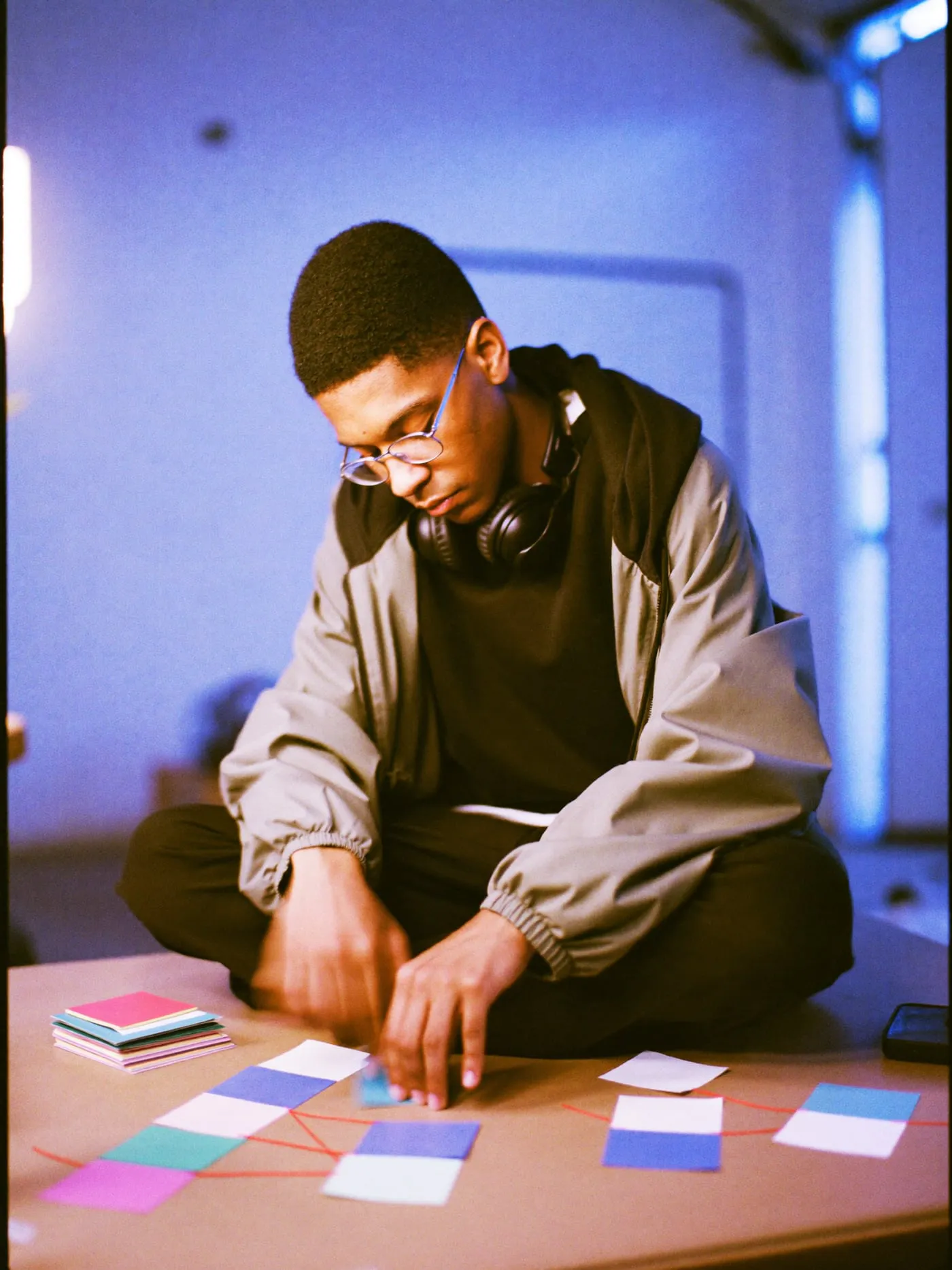 A young man in glasses sits cross legged on a desk, arranging coloured paper squares on a cork board