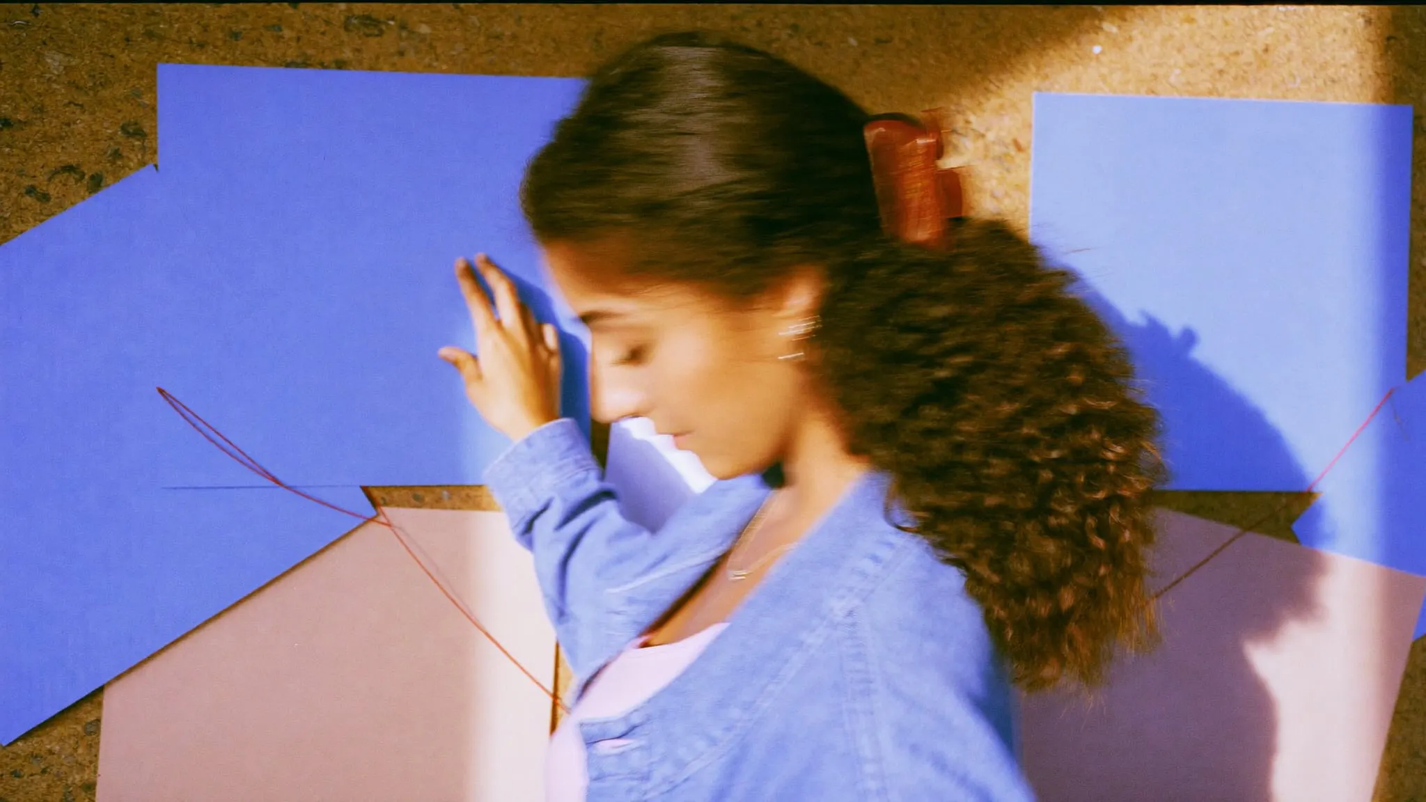 A young woman with curly hair pins periwinkle and blush paper squares into clusters on a cork wall