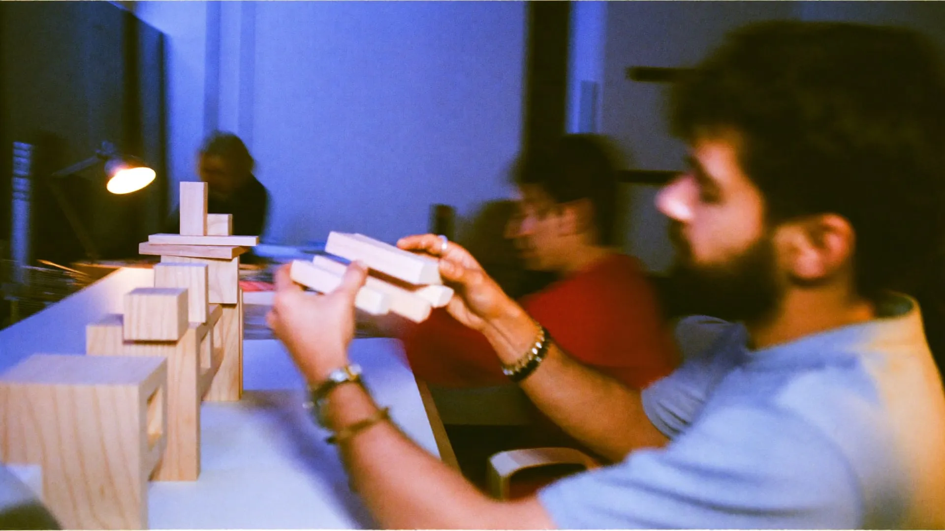 A young man stacking wooden blocks into a small model on a studio desk at night