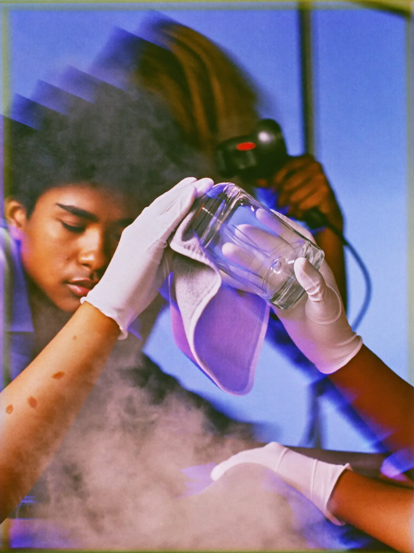 Gloved hands polishing a glass bottle while a steamer works on a shirt behind