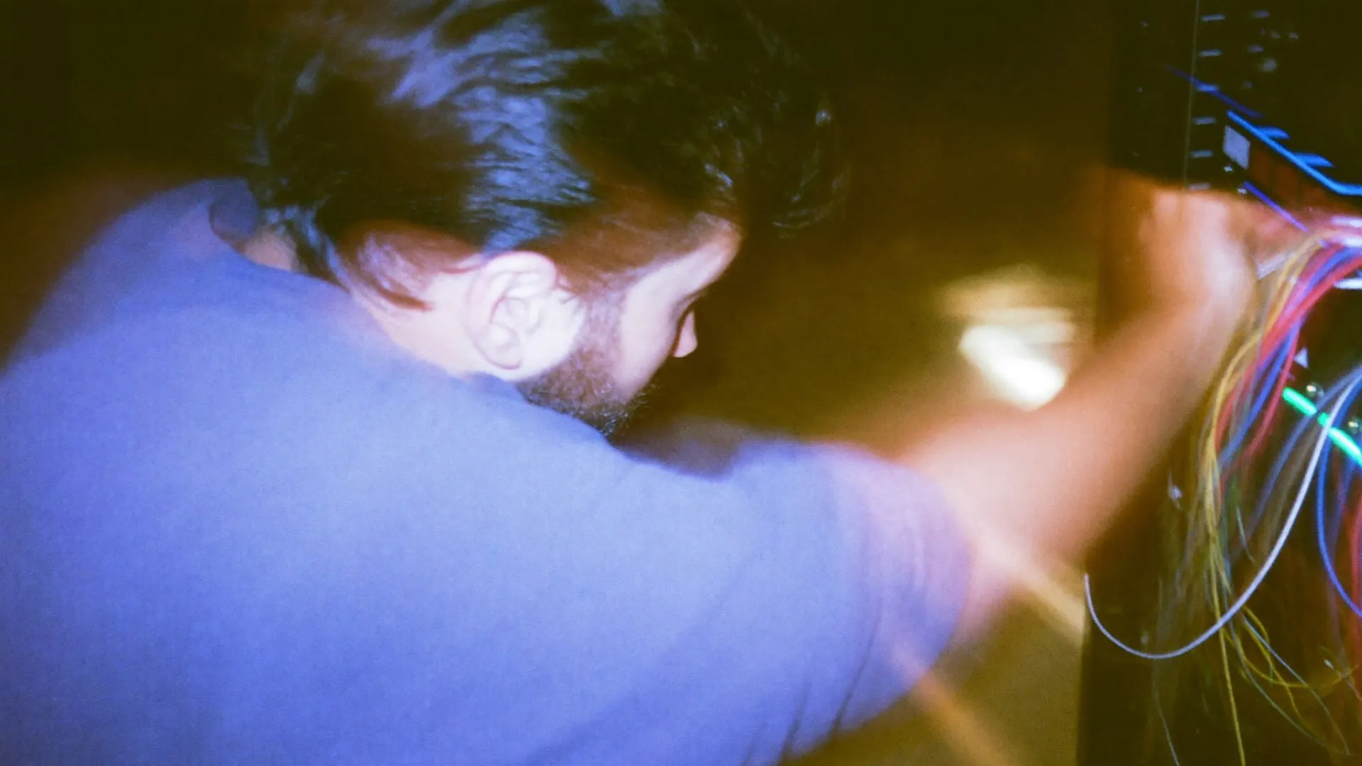 A young man seen from behind plugging cables into a server rack lit by small lights