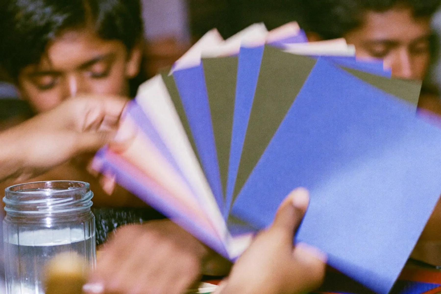 Hands fanning out squares of solid coloured card beside a brush and a jar of water