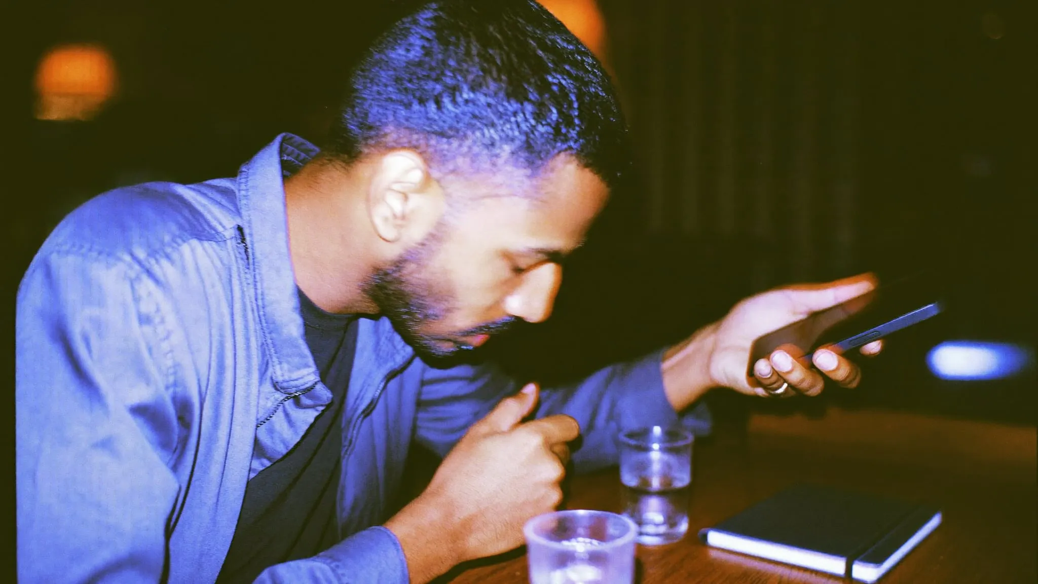 A bearded man in a cafe at night looking down at the phone in his hand