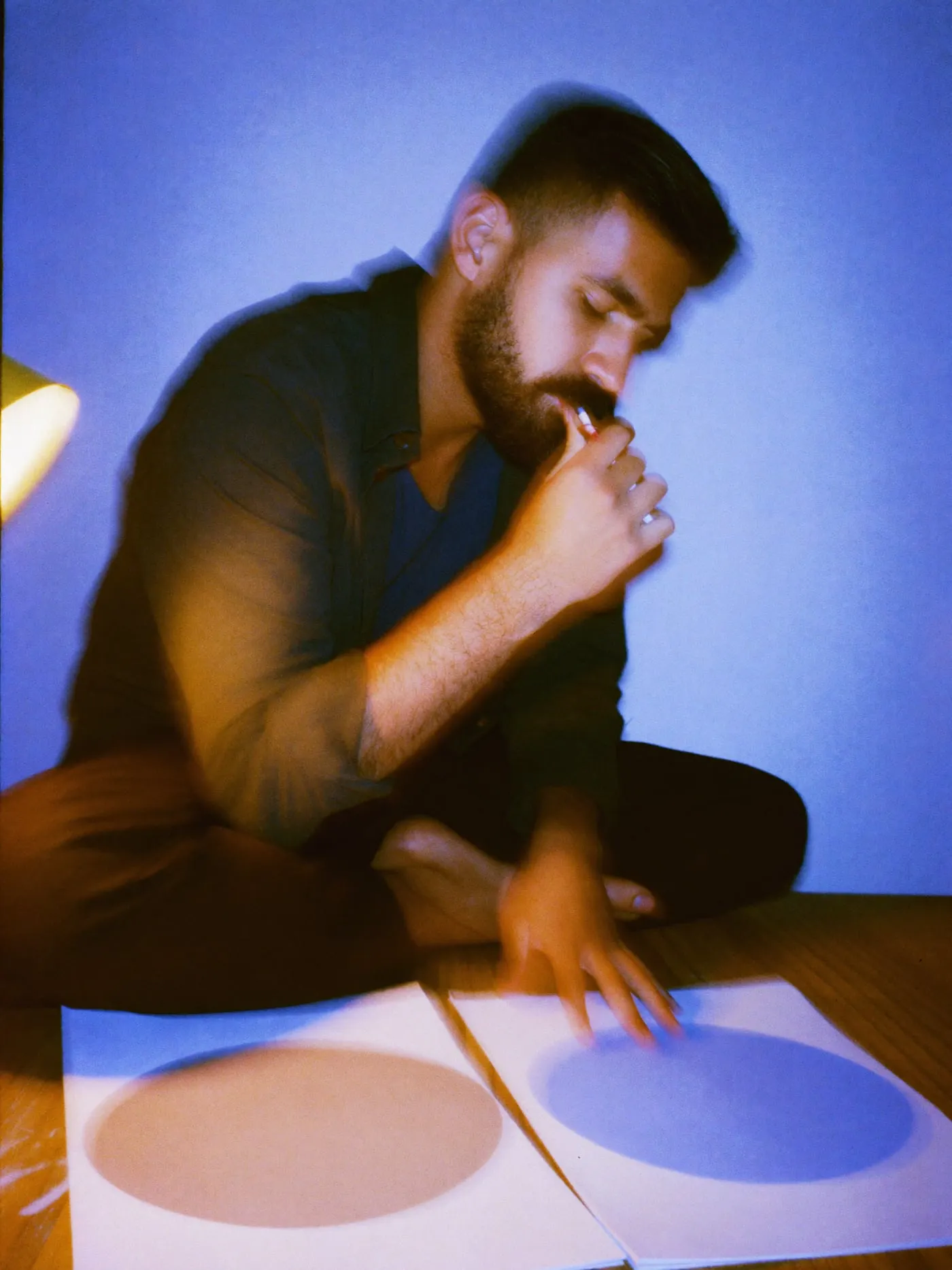 A bearded man at a studio desk at night, thinking over two painted sheets laid side by side