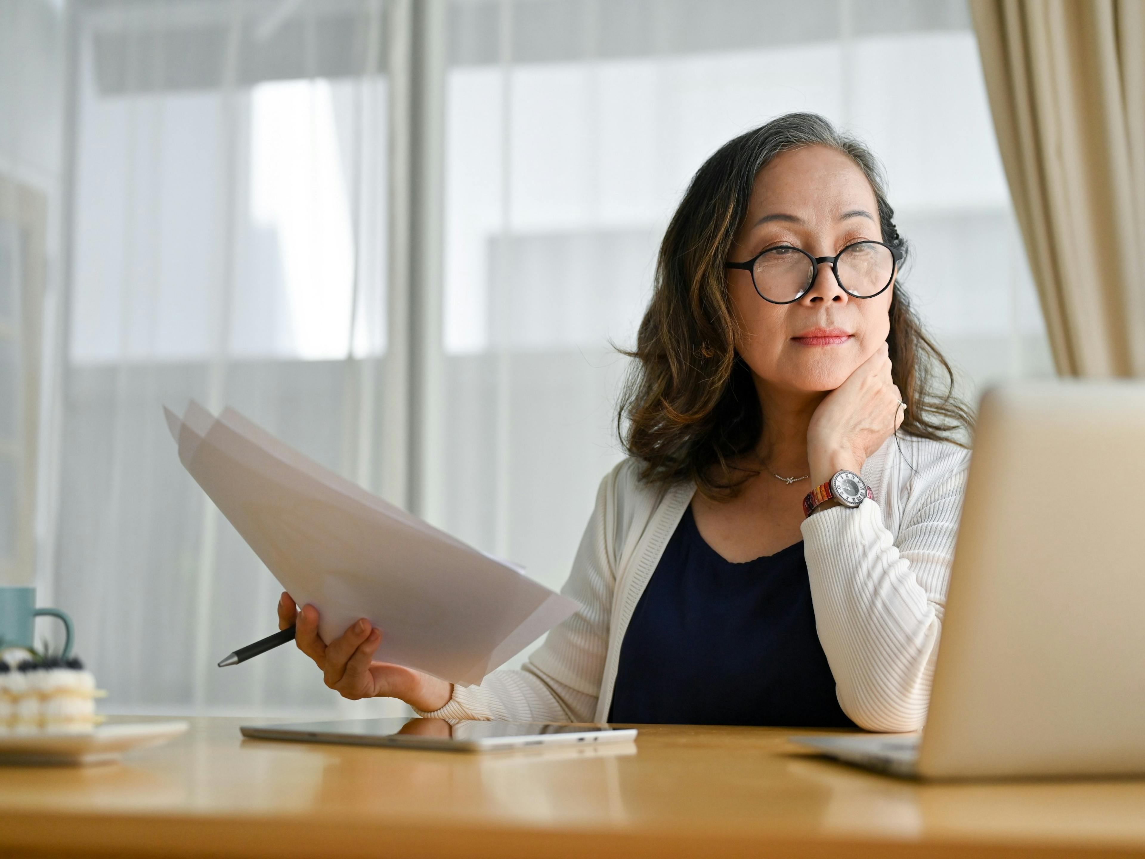 a lady with glasses working from home at a computer holding papers to review