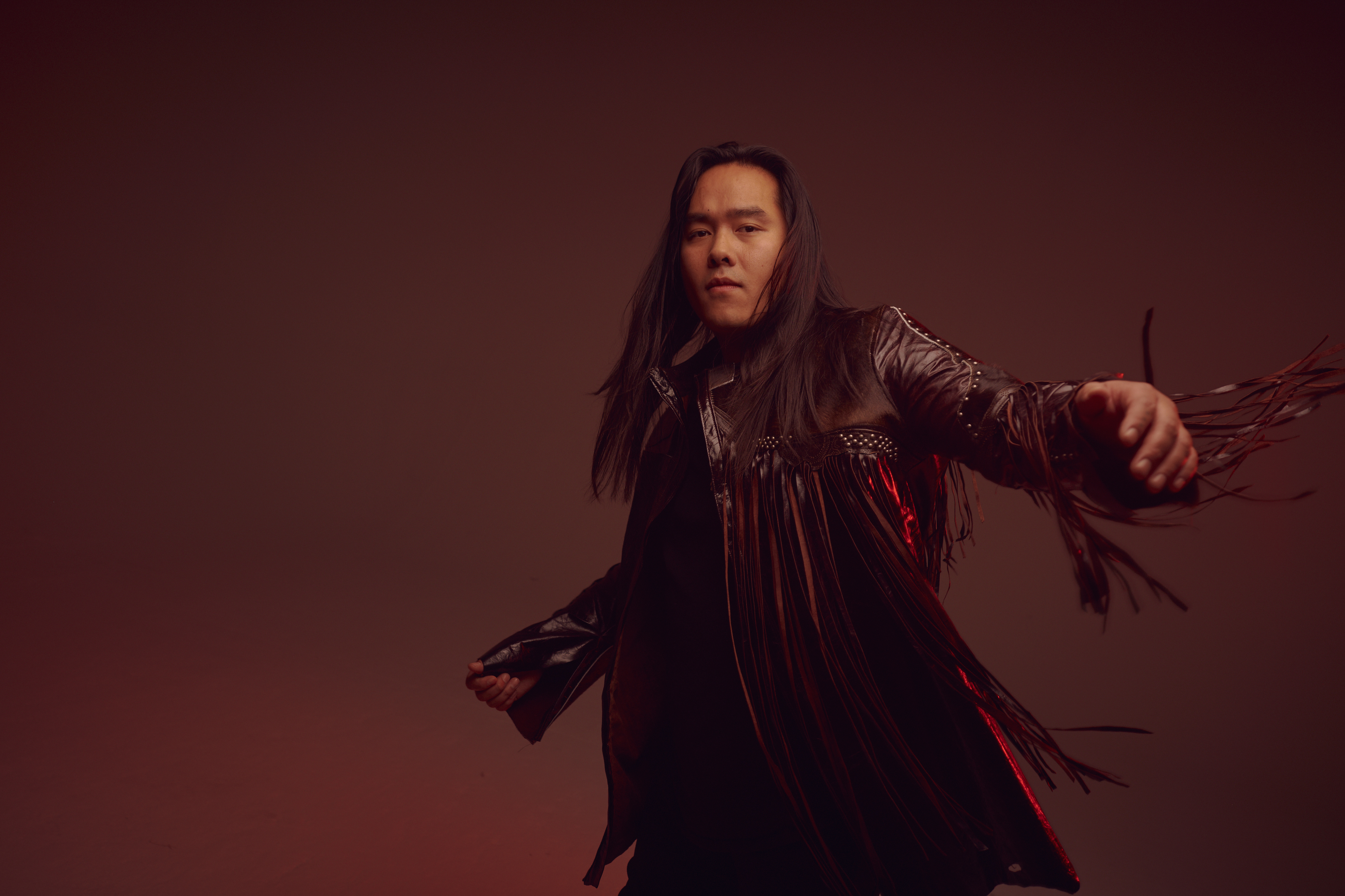 Aysanabee is pictured in his signature leather fringe trenchcoat against a brownish-red studio backdrop. He is swaying in motion. 