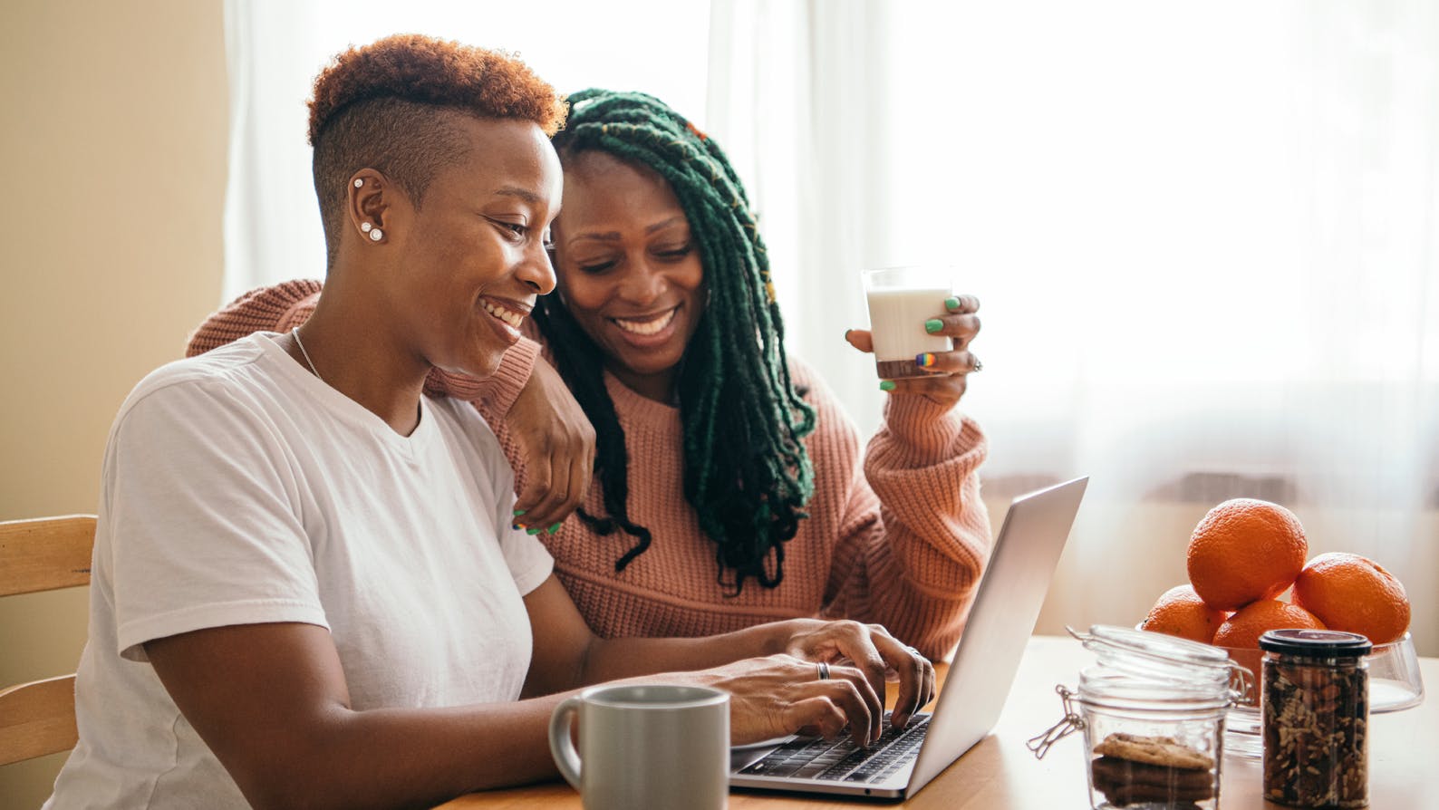 Two black women work happily on their computer at their dining table in the morning.