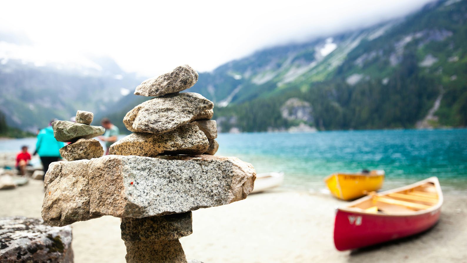 Selective focus photo of inukshuk against blue mountain lake with canoes in background