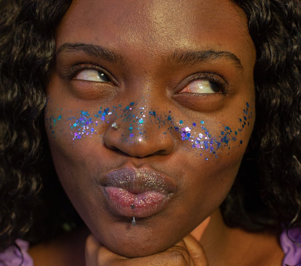 deep skin tone female model puckering lips wearing blue and purple glitter across nose and cheeks