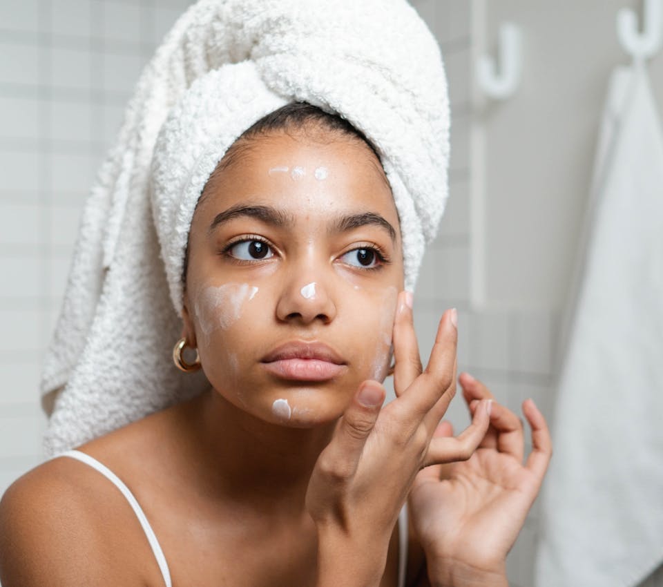 deep skin tone female model with white towel wrapped around her hair, applying lotion to face