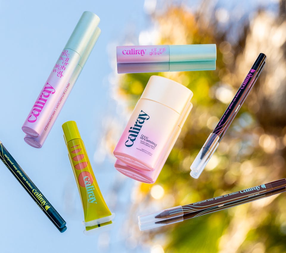 caliray clean beauty freedreaming skin tint, come hell or high water tubing mascaras in travel and regular size, surfproof eyeliner, and glazed and infused plumping lip gloss laying on mirror with reflection of tree leaves and blue sky