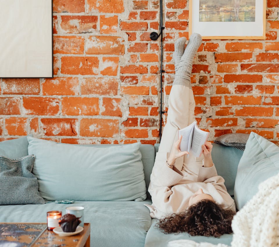female model laying on gray couch reading with her feet in the air in front of brick wall