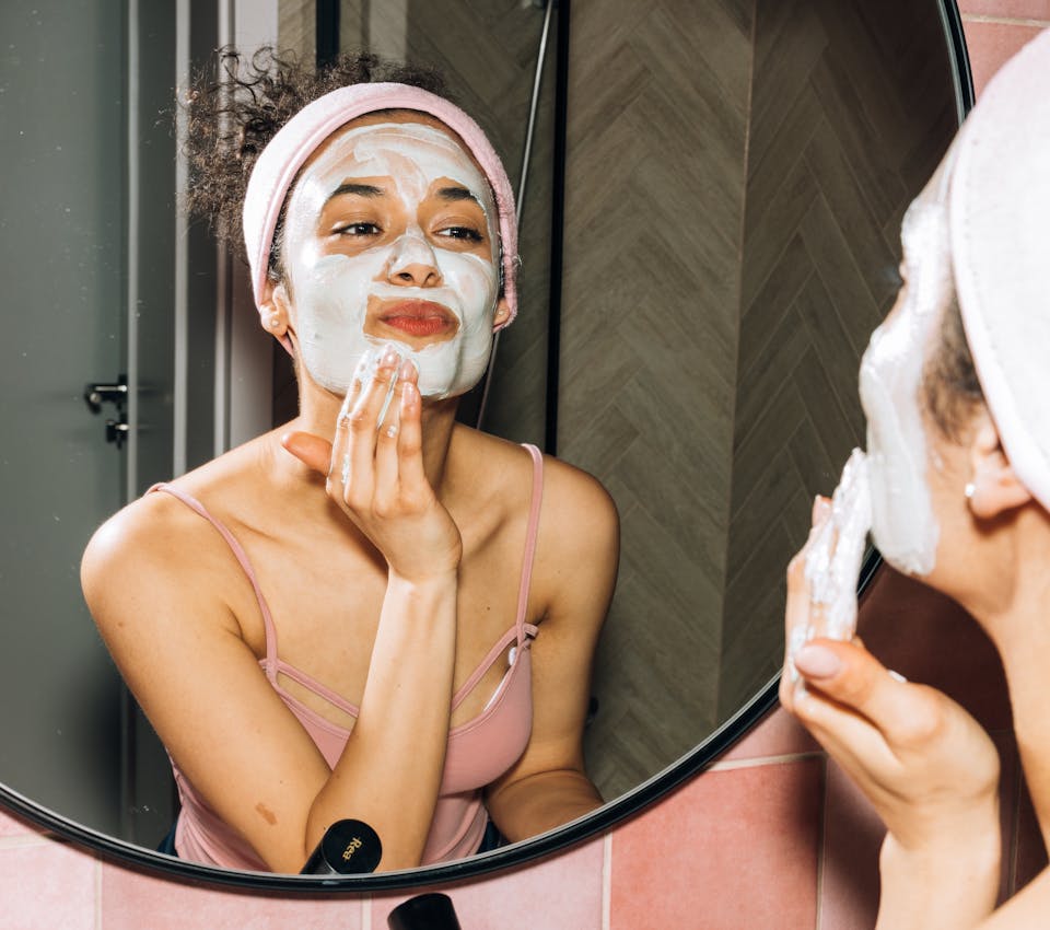 medium skin tone female model looking into bathroom mirror applying light green face mask wearing pink headband and tank top