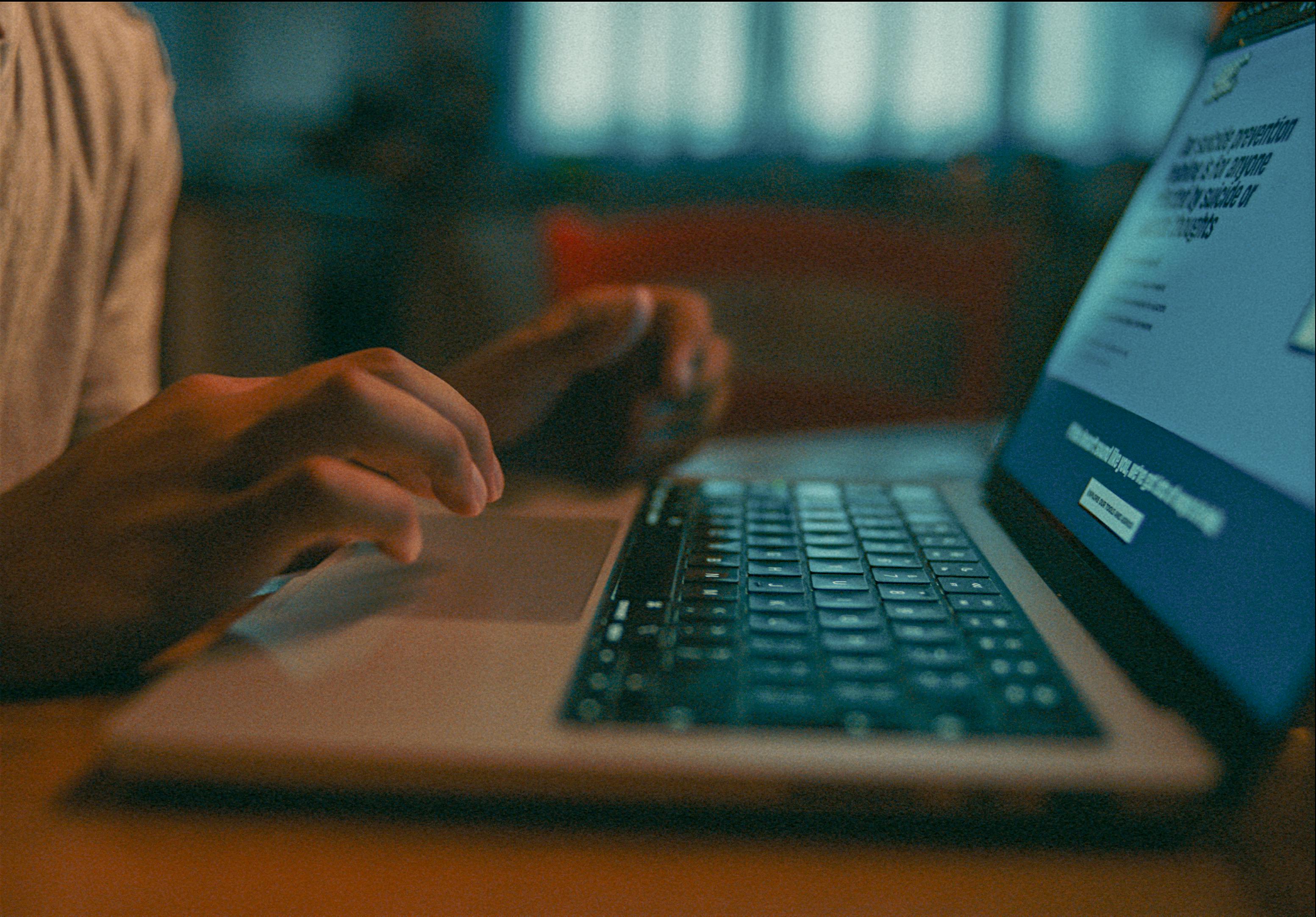 A person uses a laptop in a dimly lit room, creating a focused and intimate atmosphere. The screen glows softly as they browse the CALM website, highlighting their fingers on the keyboard.