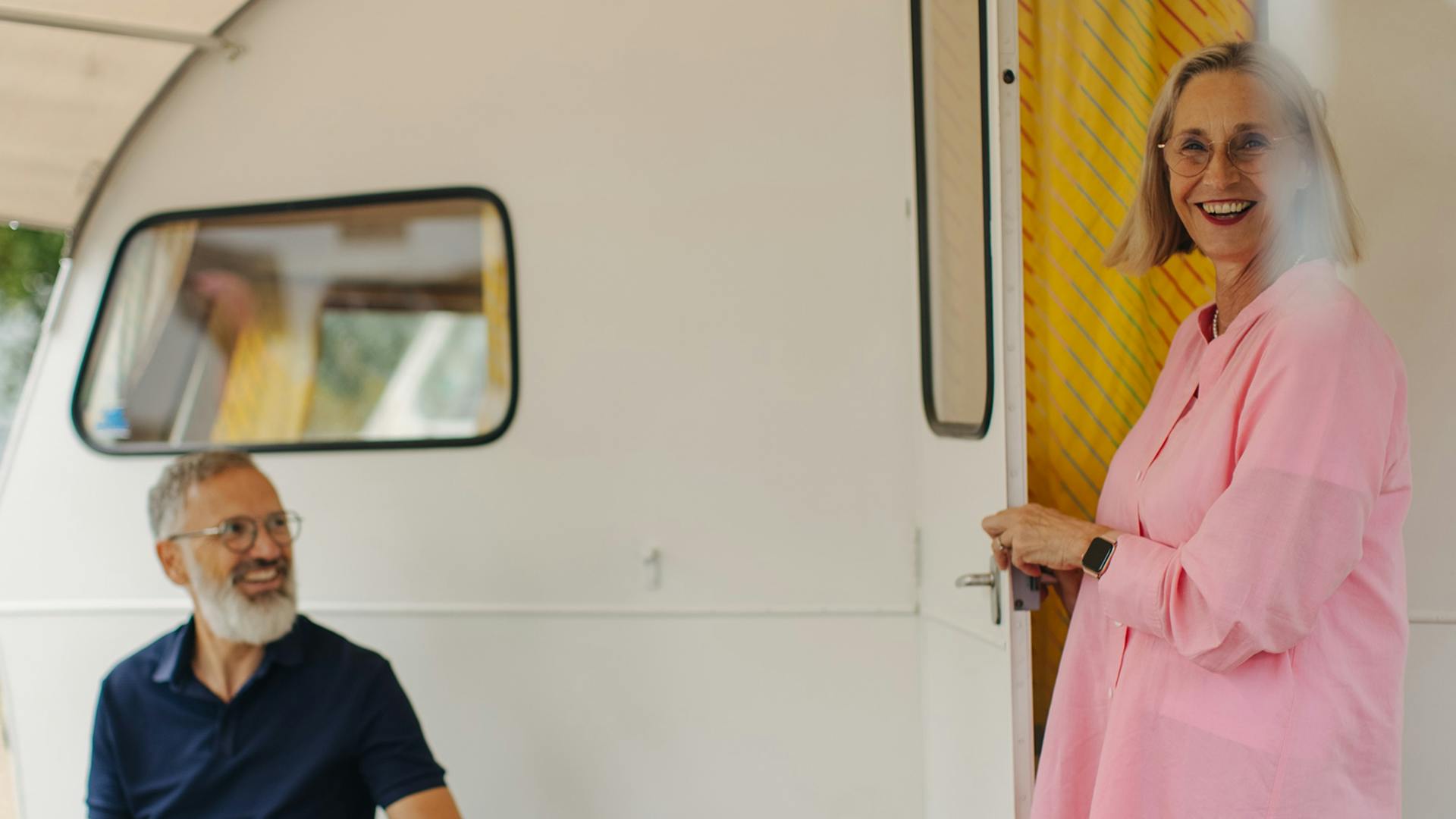 Frau kommt aus einem kleinen Wohnwagen während Mann davor sitzt