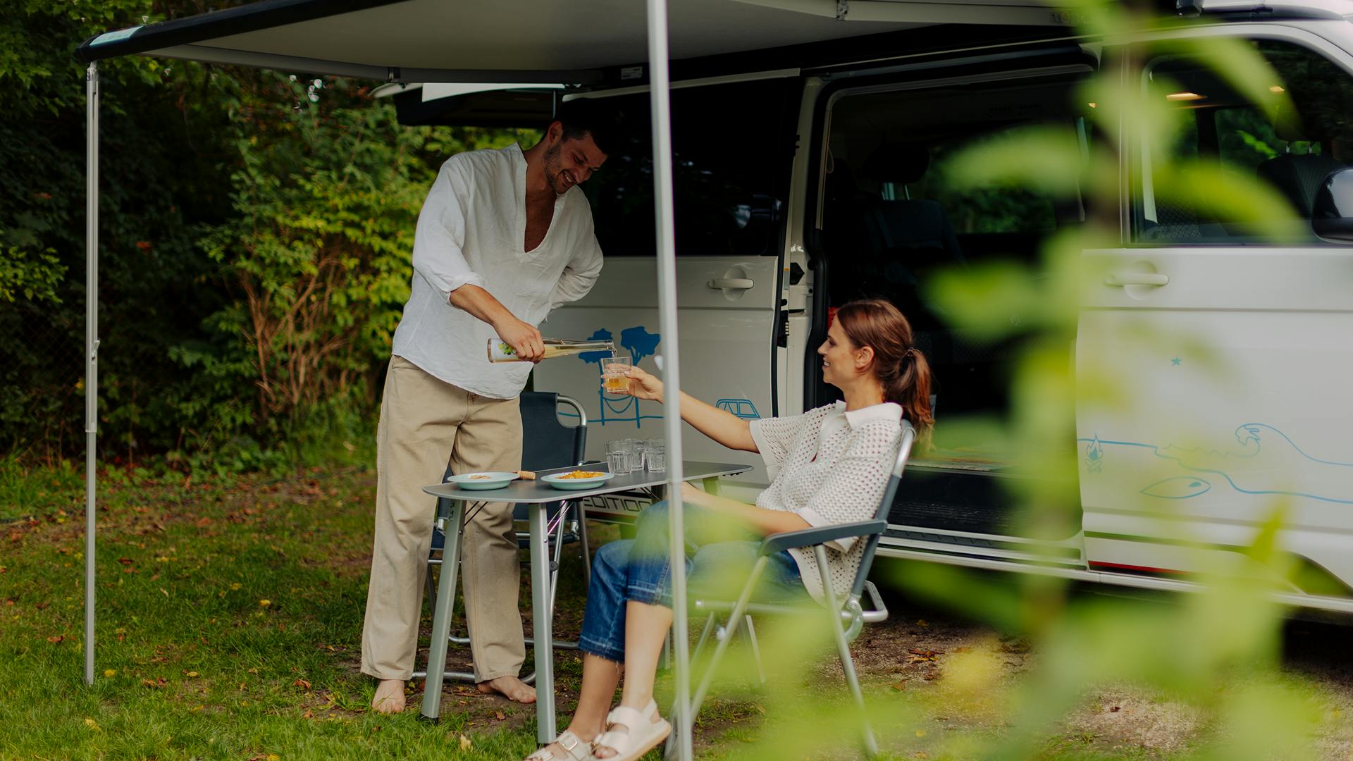 Mann steht vor einem Volkswagen California und schenkt Frau Getränk ein während sie sitzt