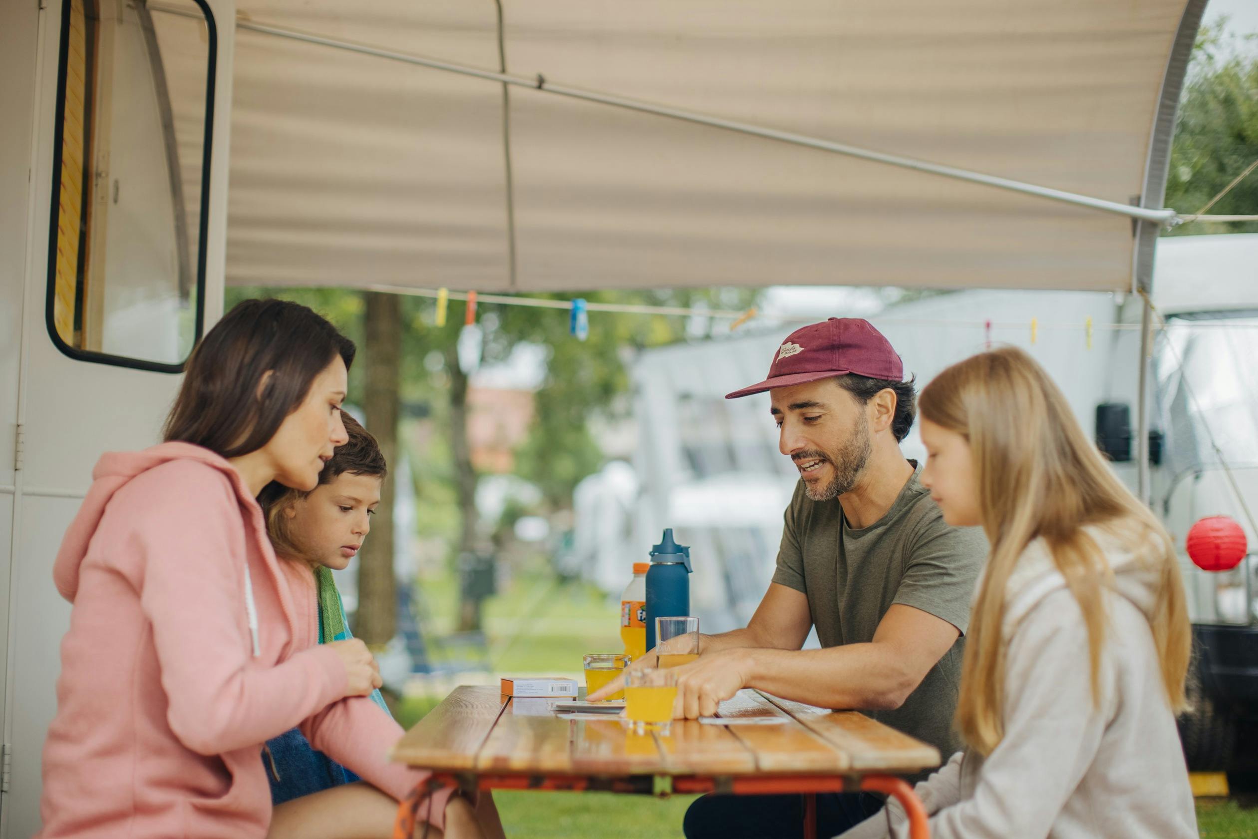 Familie mit zwei Kindern an einem Campingtisch unter der Markise eines Wohnwagens