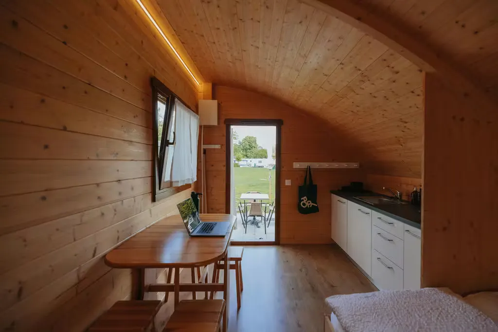 Vista interior de la Beach Cabin en el camping Erlach: vista desde la cama, con una mesa y sillas a un lado y una cocina americana al otro; la puerta de la cabaña da a la terraza.