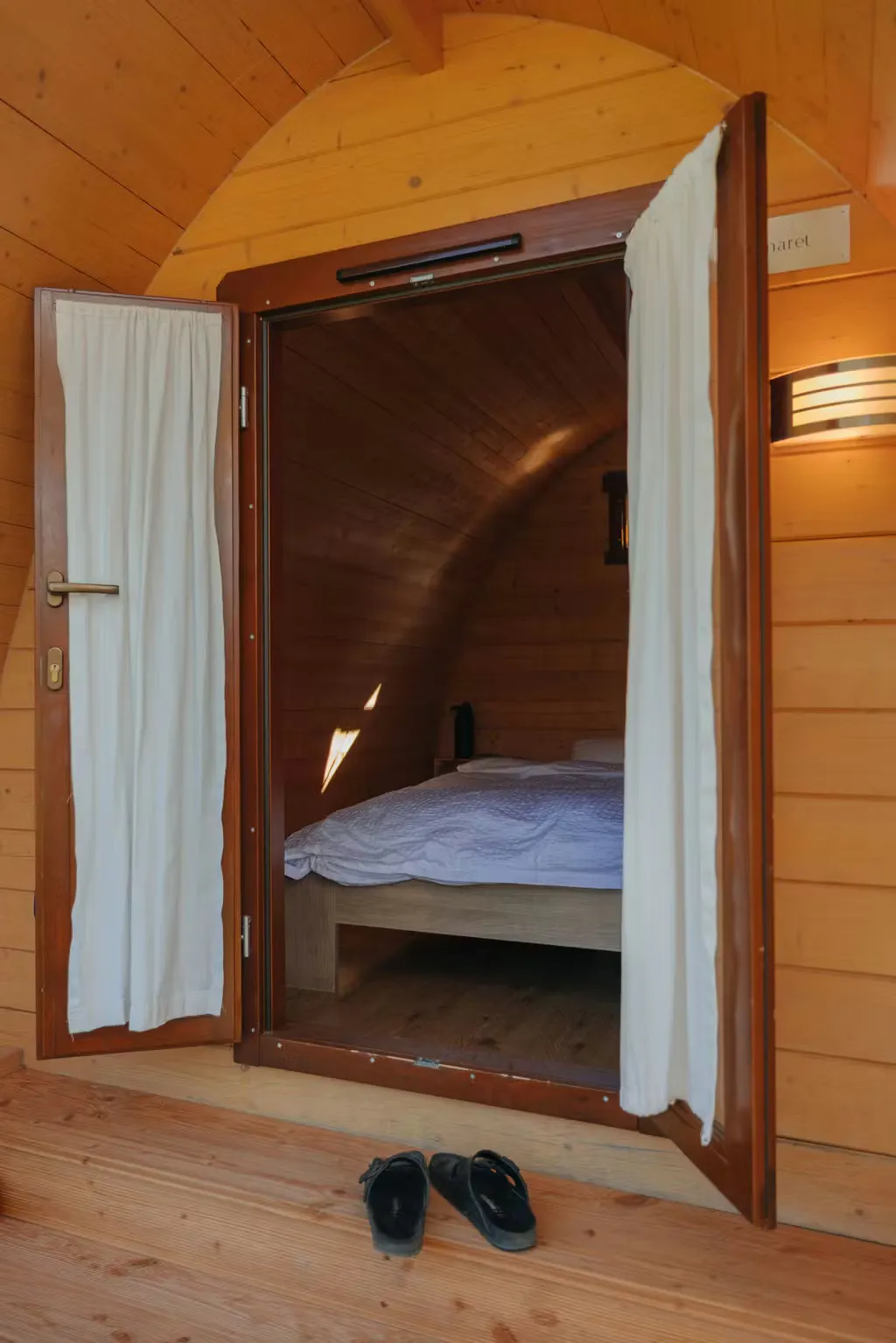 Türen und Fenster des Beach Hut auf dem Camping Erlach stehen offen, die Vorhänge sind zugezogen, ein Doppelbett steht drin, ein Paar Schuhe stehen draussen