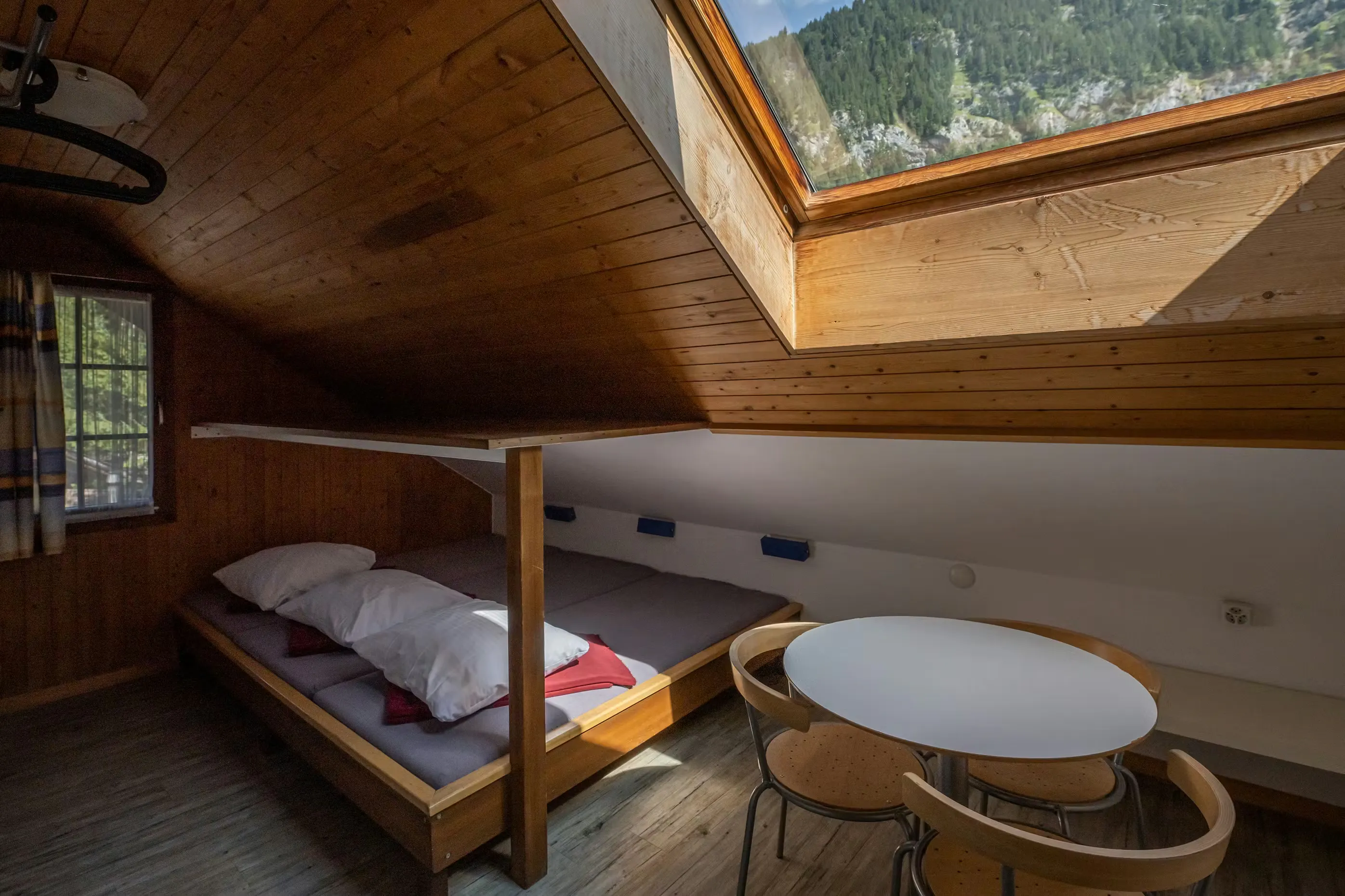 Herbergszimmer mit drei Betten und einem Tisch mit drei Stühlen, Dachfenster mit Blick auf die Berge und die hereinscheinende Sonne