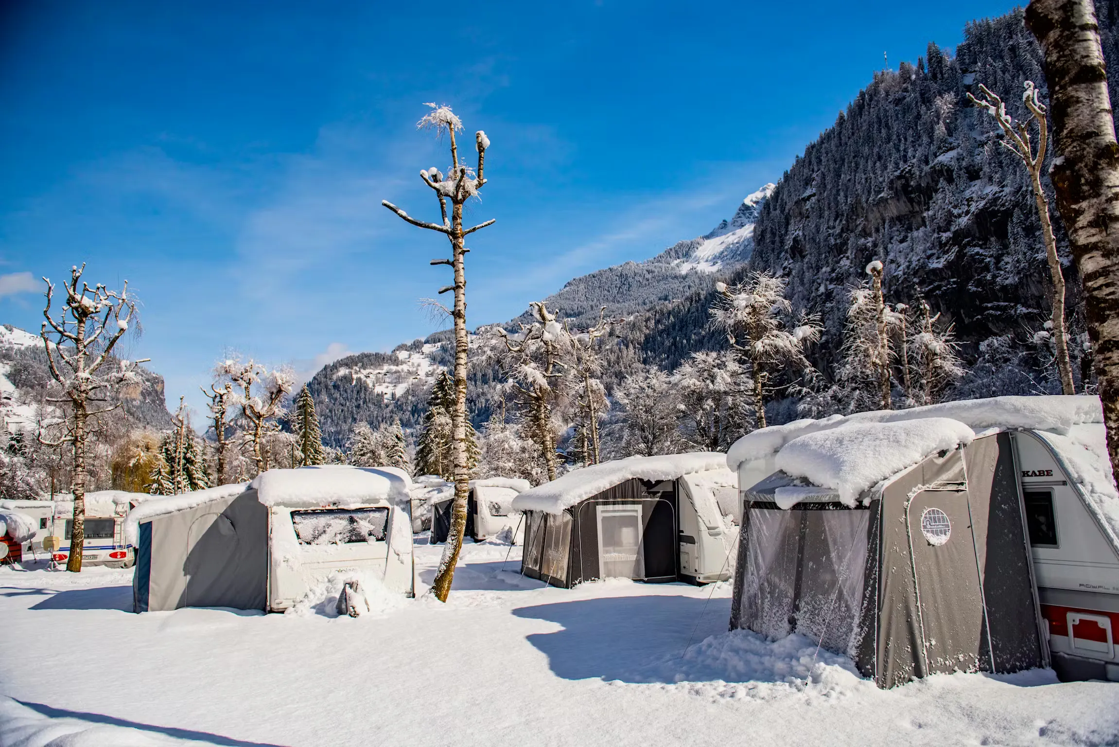 mehrere Wohnwagen mit Vorzelten auf einem verschneiten Campingplatz mit blauem Himmel und Bergen im Hintergrund