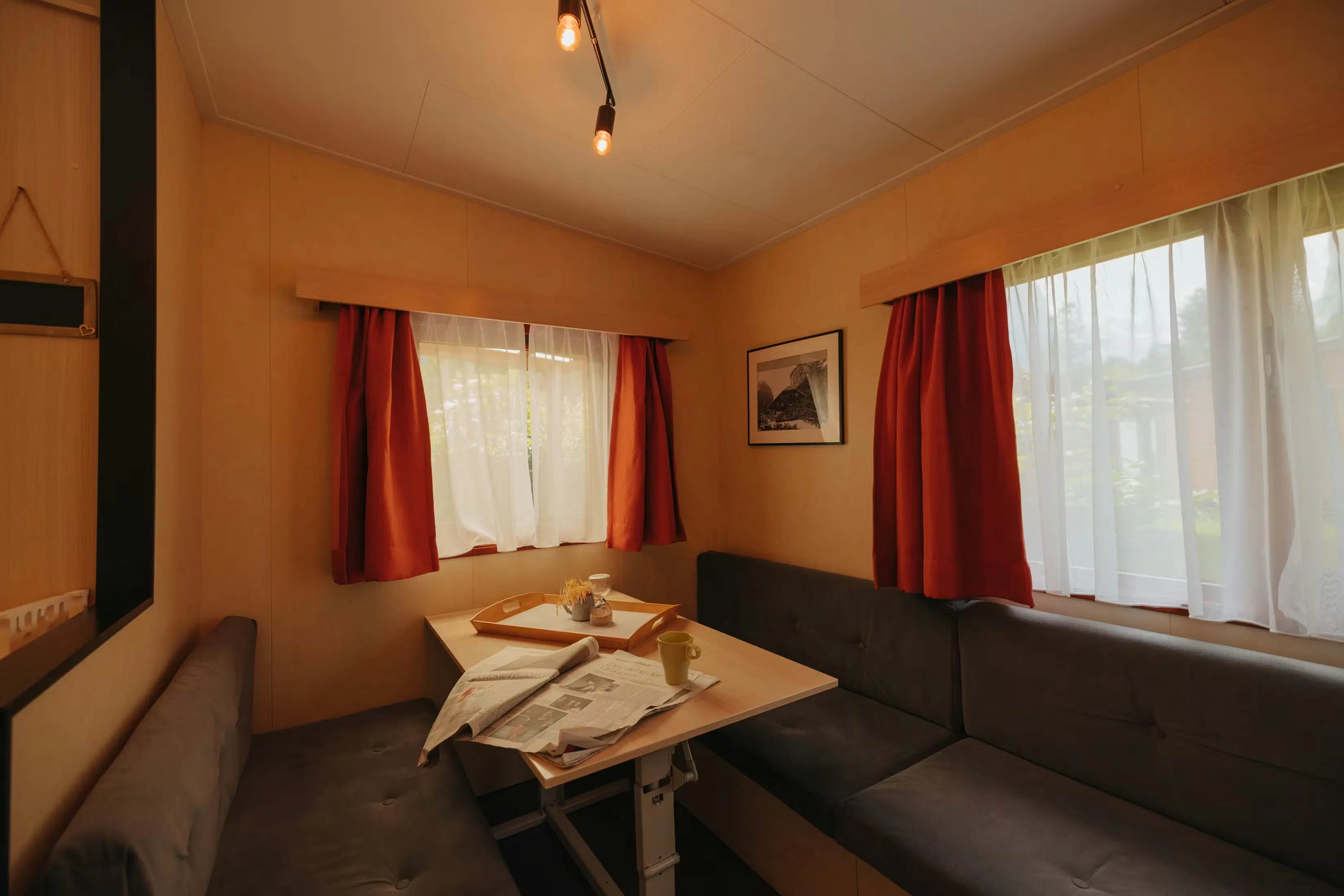 Ecktisch mit Bänken mit grauen Bezügen, mehrere Fenster und rote Vorhänge. Zeitung und Becher auf dem Tisch.