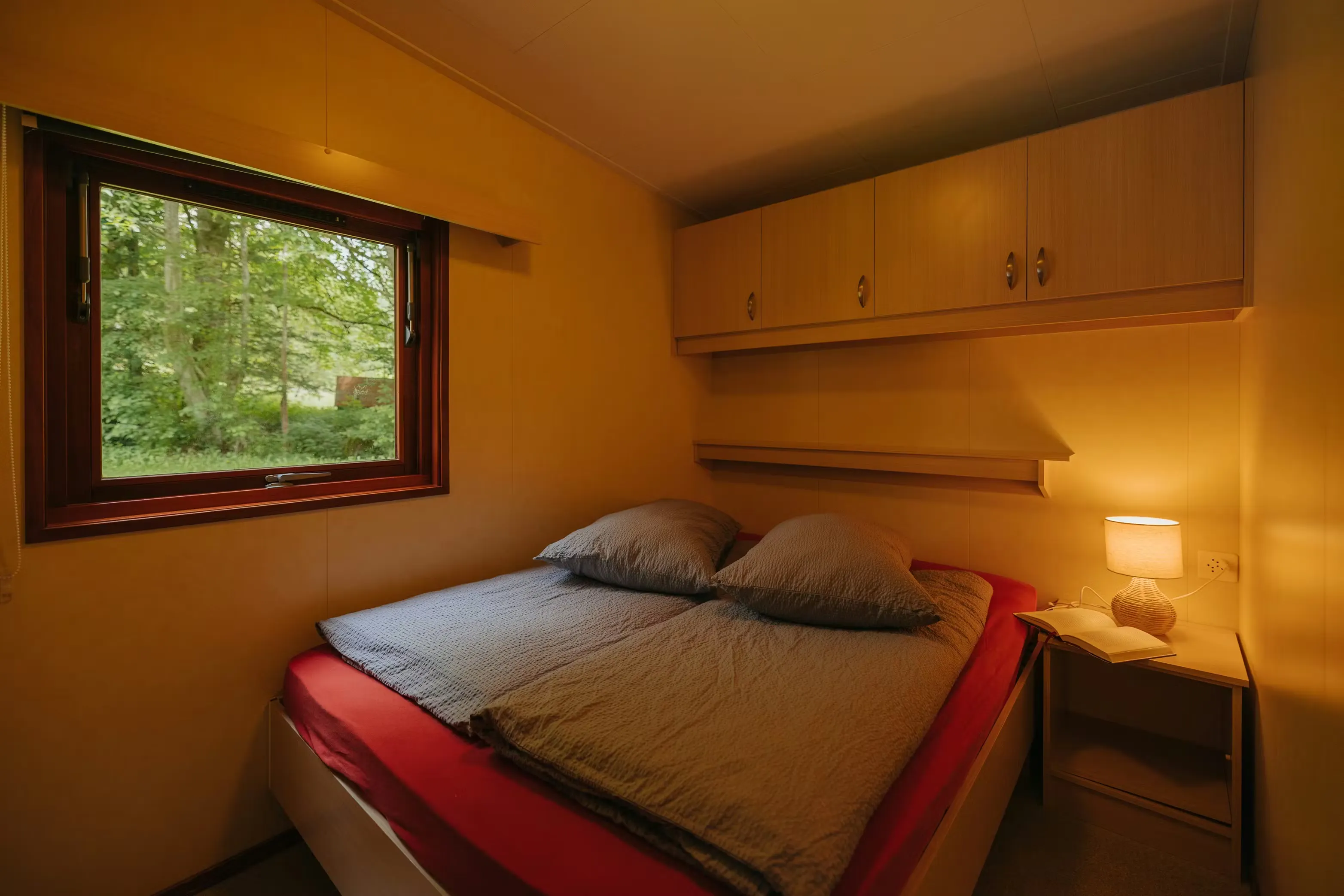 Schlafzimmer des Mobilheims Jungfrau mit einem Doppelbett, einem Nachttisch mit Beleuchtung, Schränken über dem Bett und einem Fenster an der Seite des Zimmers