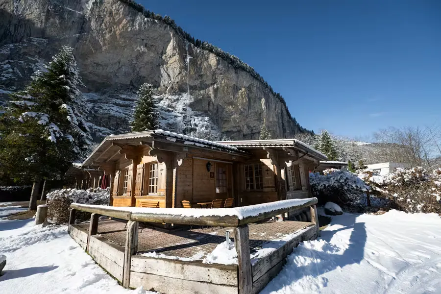 Bungalow im Winter mit schneebedecktem Boden und einem Wasserfall, der von den Felswänden im Hintergrund herabstürzt