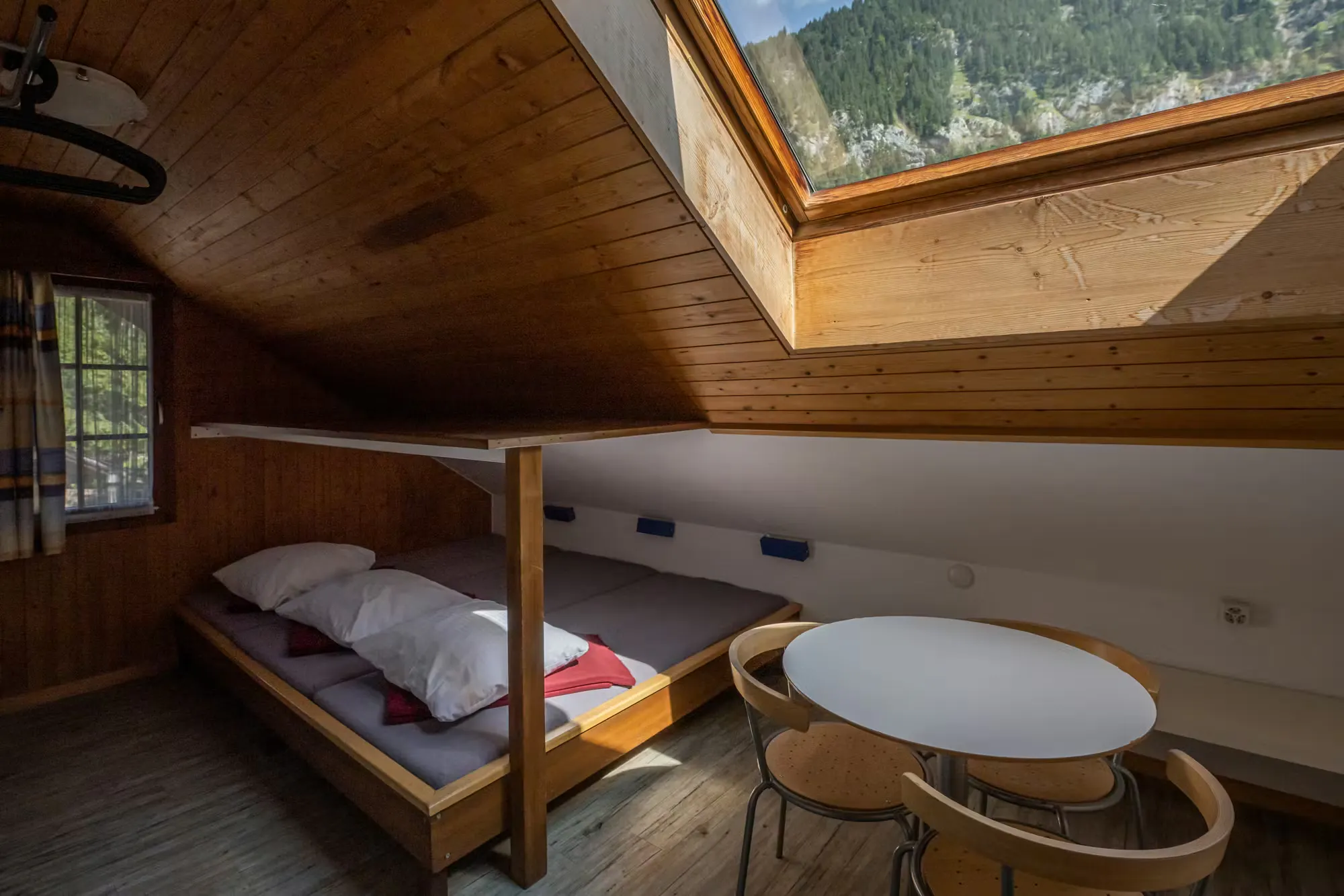 Herbergszimmer mit drei Betten und einem Tisch mit drei Stühlen, Dachfenster mit Blick auf die Berge und die hereinscheinende Sonne