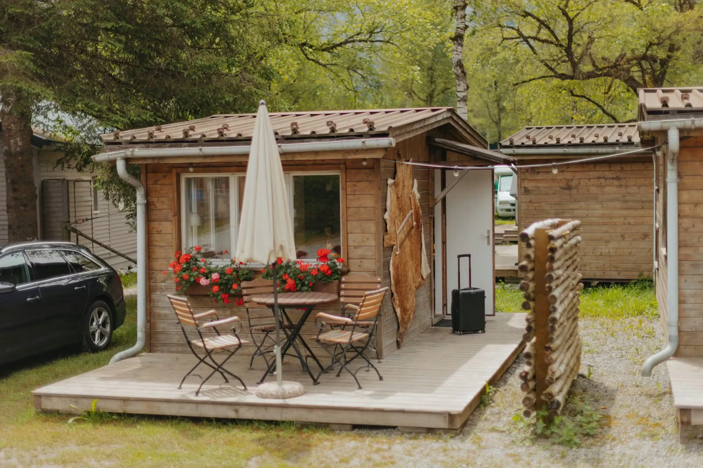 Mietunterkunft Eigerhütte mit einem Koffer vor der offenen Tür und einem Tisch und vier Stühlen auf der Terrasse. Der Sonnenschirm ist geschlossen, aber unter dem Fenster blühen schöne rote Blumen