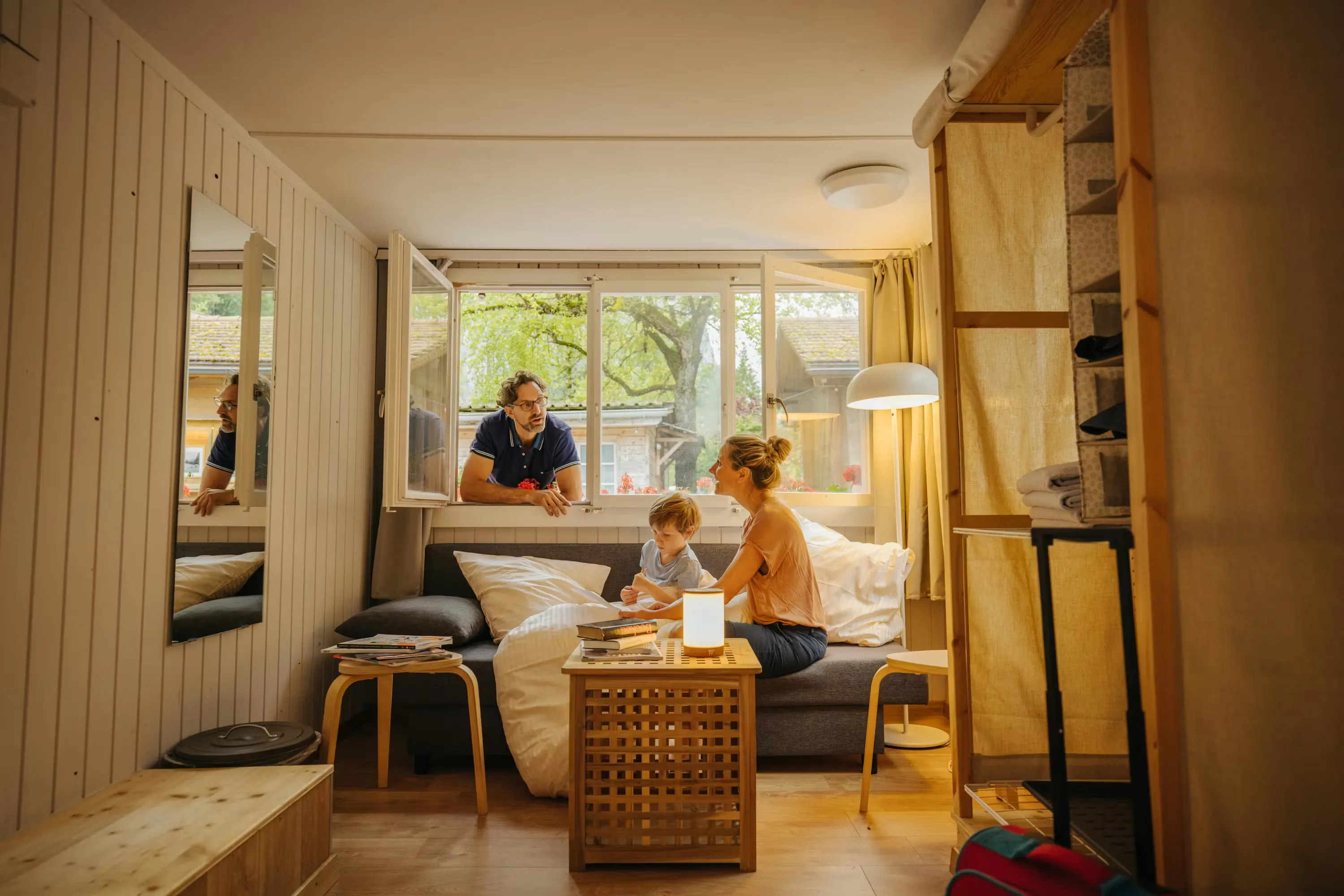 Mutter und Kind sitzen auf dem Sofa der Eigerhütte und der Vater schaut durch das offene Fenster von der Terrasse ins Innere