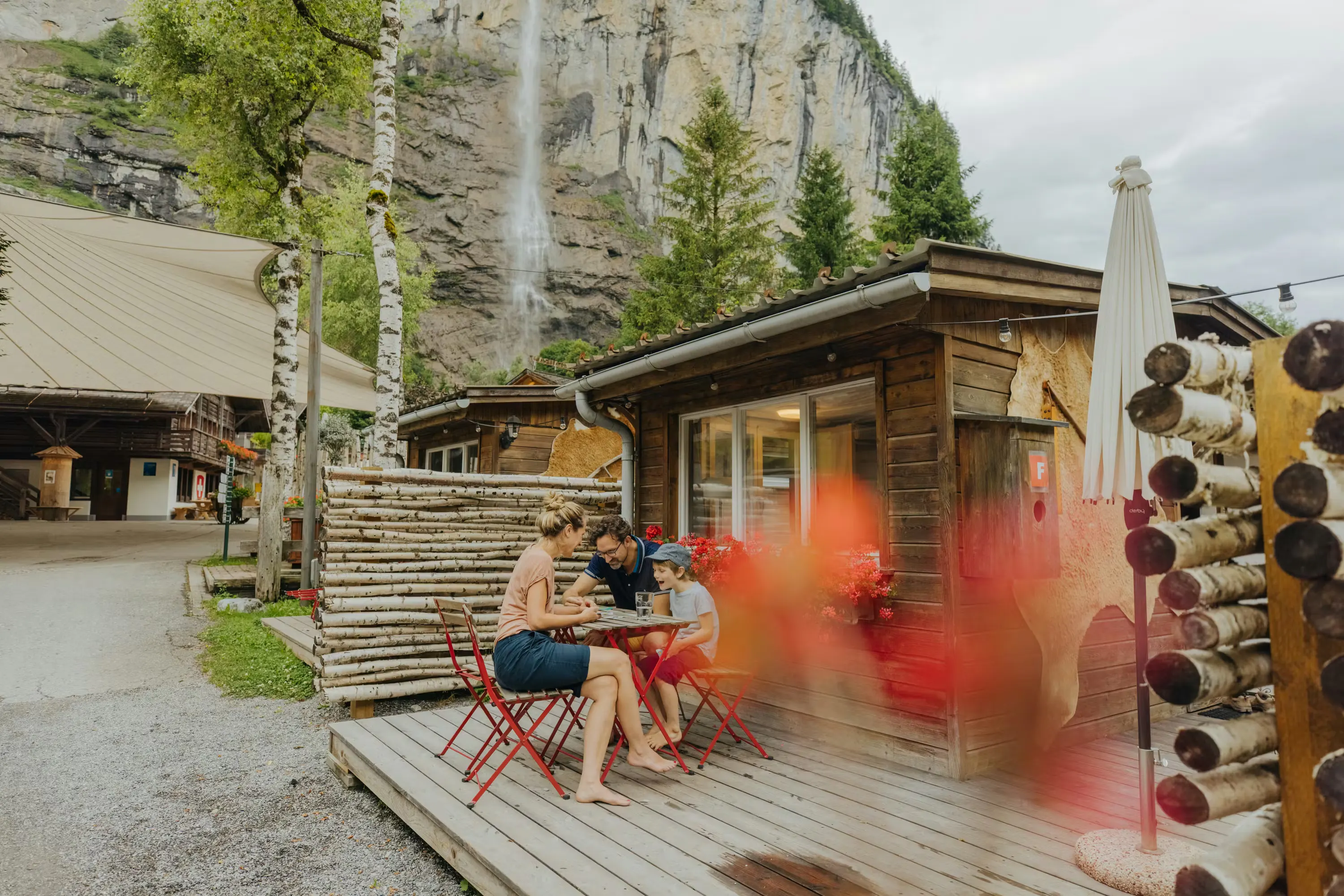 zwei Erwachsene und ein Kind auf der Terrasse der Eigerhütte des Camping Jungfrau mit dem Staubbach-Wasserfall im Hintergrund